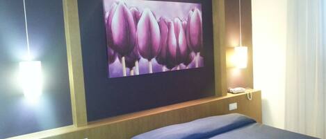 Una habitación de hotel con una cama, un televisor de pared mostrando flores moradas, una lámpara de noche y un teléfono.