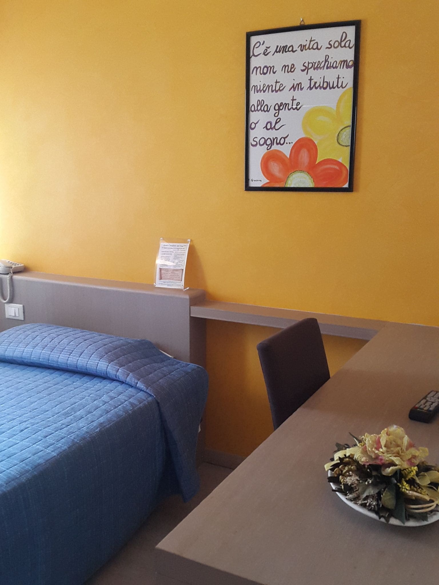A hotel room with a bed, a table with a plate of flowers, and a wall with a framed quote.