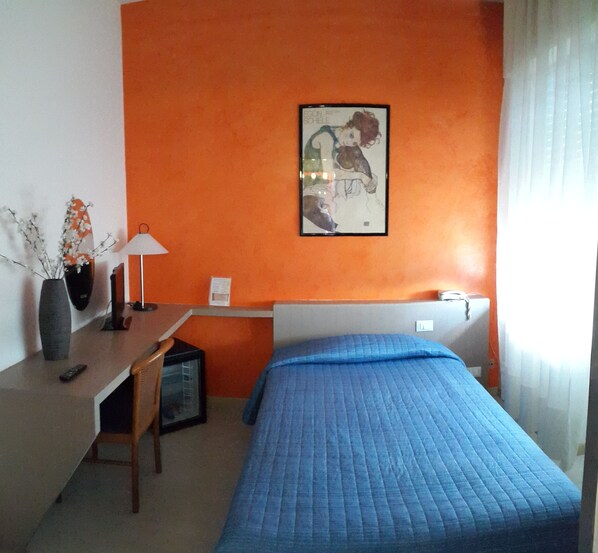 A bedroom with a bed, a desk, a chair, a lamp, and a framed picture on the wall.