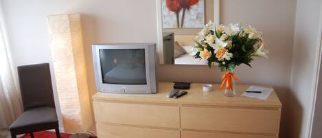 Habitación de hotel con tocador de madera, televisión, sillón, un jarrón de flores y un cuadro en la pared.