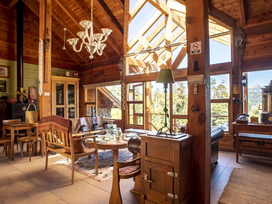 Interior rústico de cabana com vigas de madeira, um candelabro, uma janela grande e um fogão antigo.