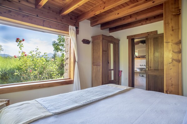 A bedroom with a large window, wooden ceiling beams, a bed with a white mattress, a wooden wardrobe, and a view of greenery and mountains. - Colina del Manzano (Villa La Angostura)