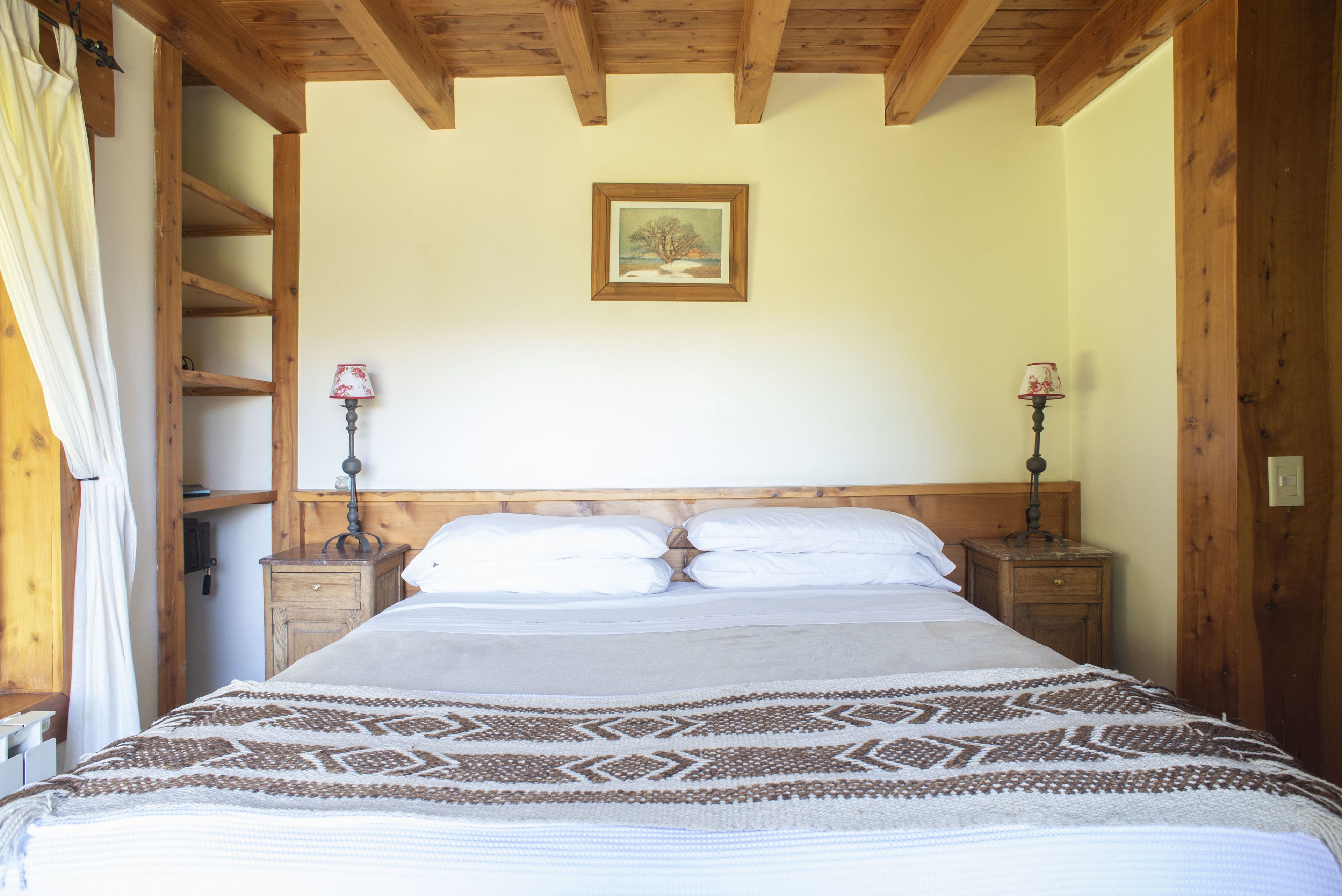 A bedroom with a wooden ceiling, a bed with white linens, two bedside tables with lamps, and a painting on the wall.