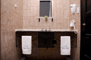 Une salle de bain avec un mur recouvert de carreaux beiges, une étagère en verre au-dessus du lavabo et un sèche-cheveux fixé au mur.