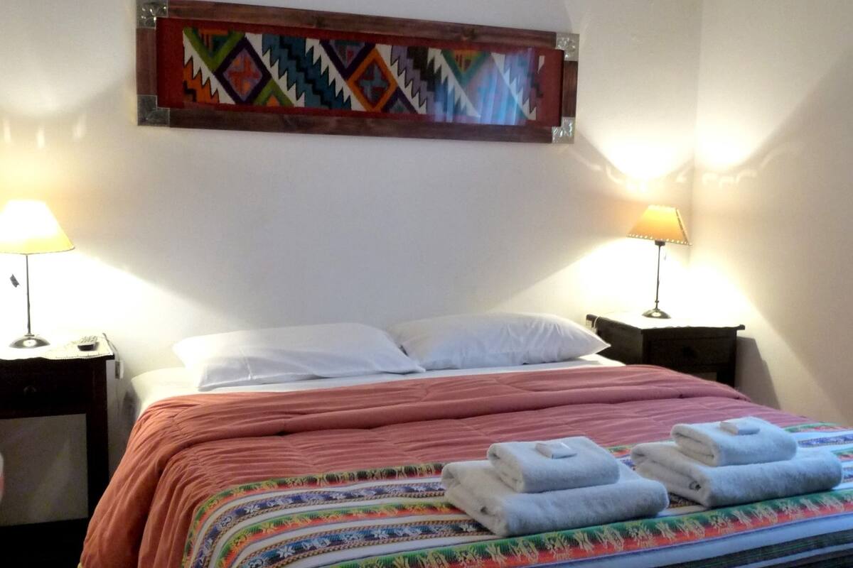 Ein ordentlich bezogenes Bett mit farbenfroher Tagesdecke, zwei Nachttischlampen und einem dekorativen Wandbild.