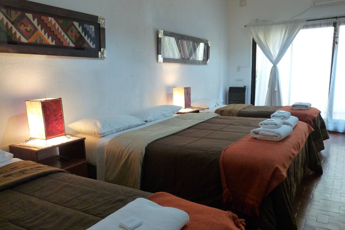 Ein Hotelzimmer mit drei Betten, jedes mit einer andersfarbigen Decke, einem Nachttisch mit Lampe und einem Gemälde an der Wand.