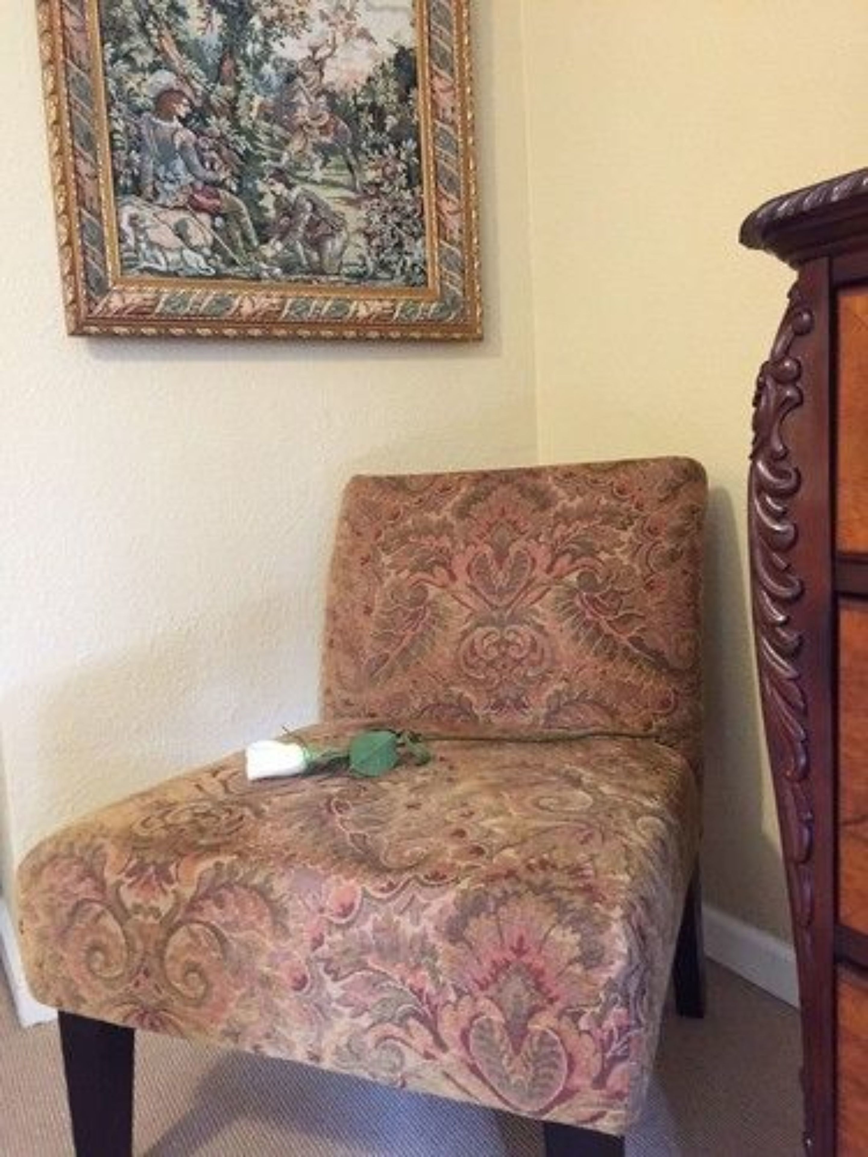 A chair with a patterned fabric, a framed painting on the wall, and a wooden cabinet.