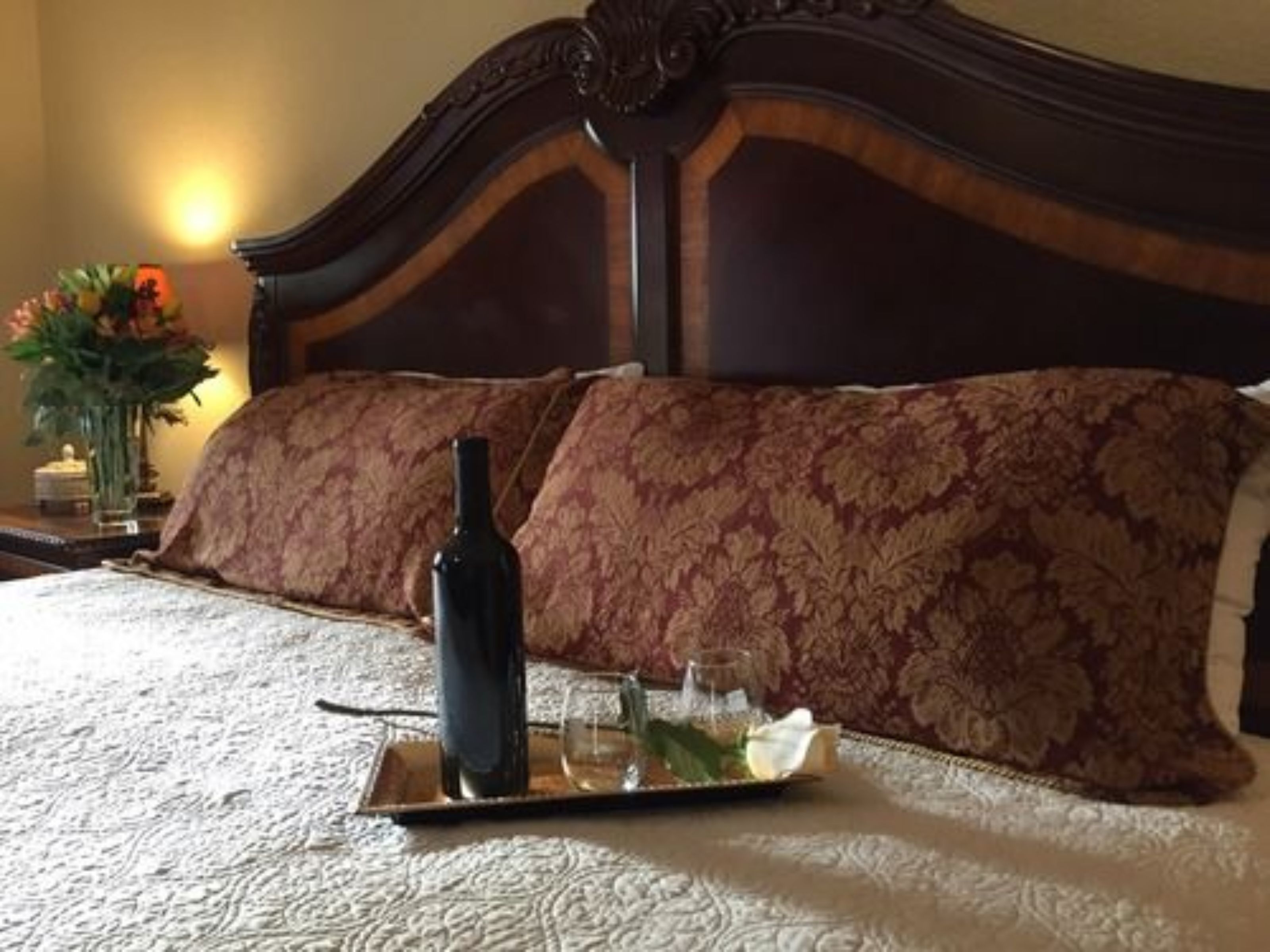 A neatly made bed with a tray holding a bottle, glasses, and a flower.