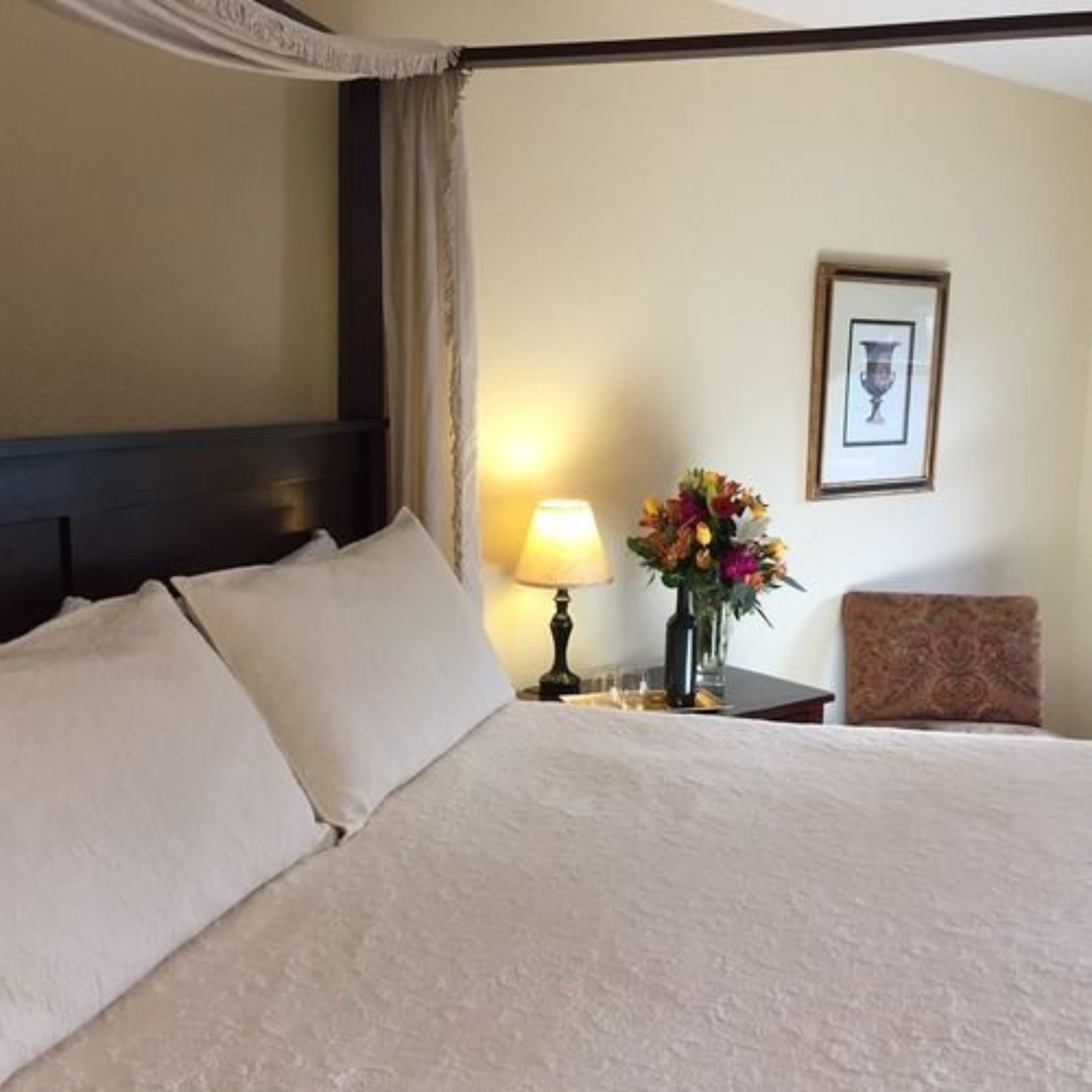 A hotel room with a large bed, a nightstand with a lamp and a vase of flowers, a framed picture on the wall, and a chair.