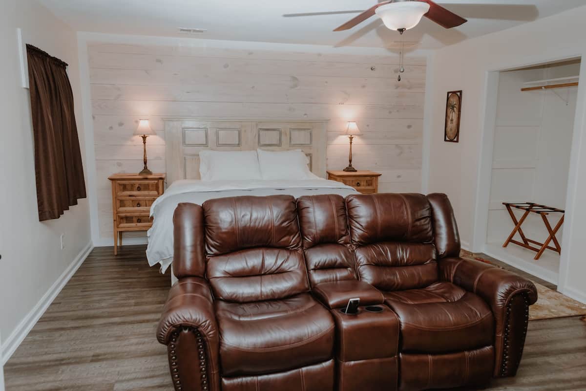 A bedroom with a large bed, two bedside tables with lamps, a ceiling fan, and a brown leather sofa.