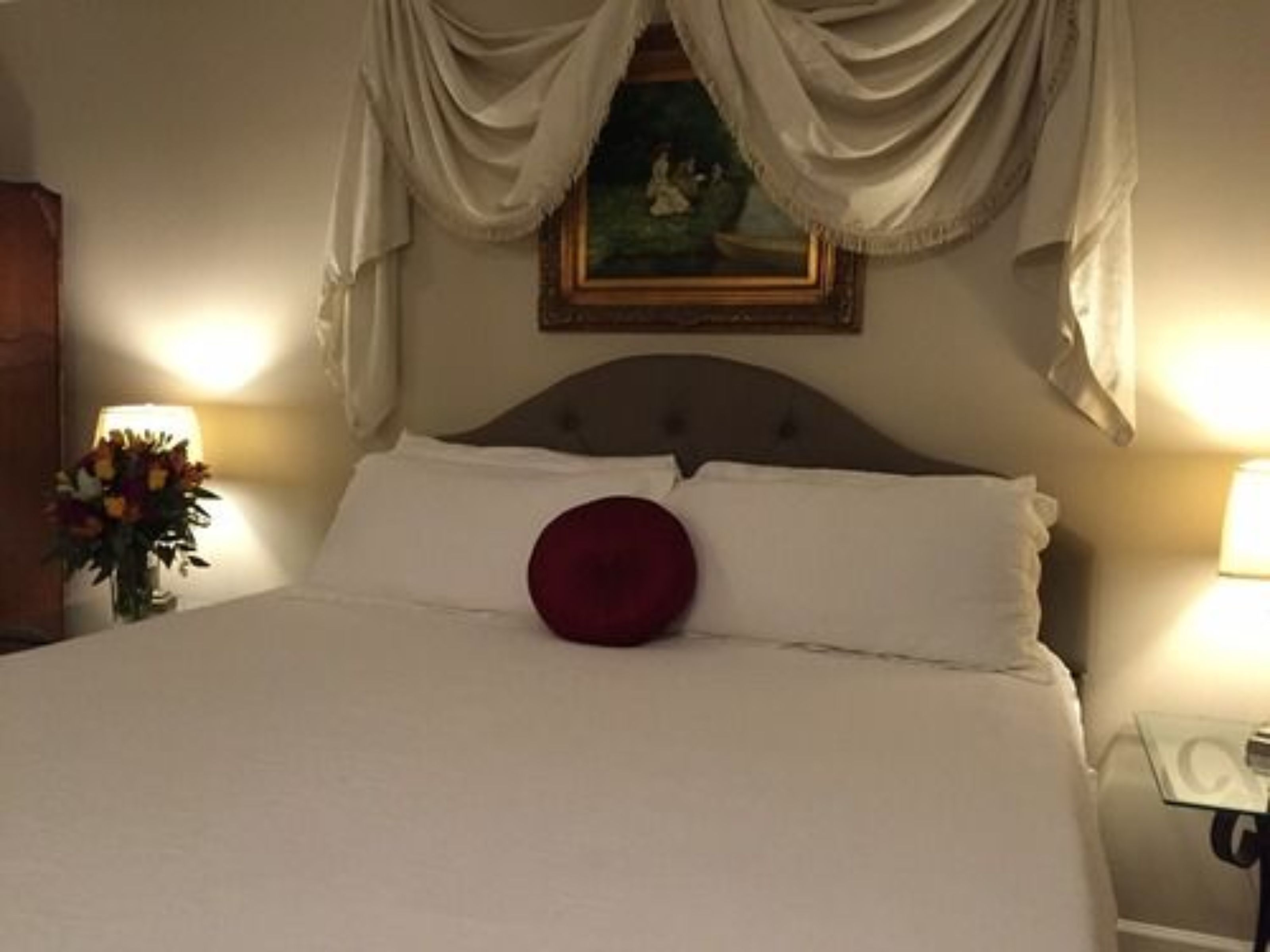 Een net opgemaakt bed met witte lakens en één rood kussen, een schilderij erboven en twee bedlampen.