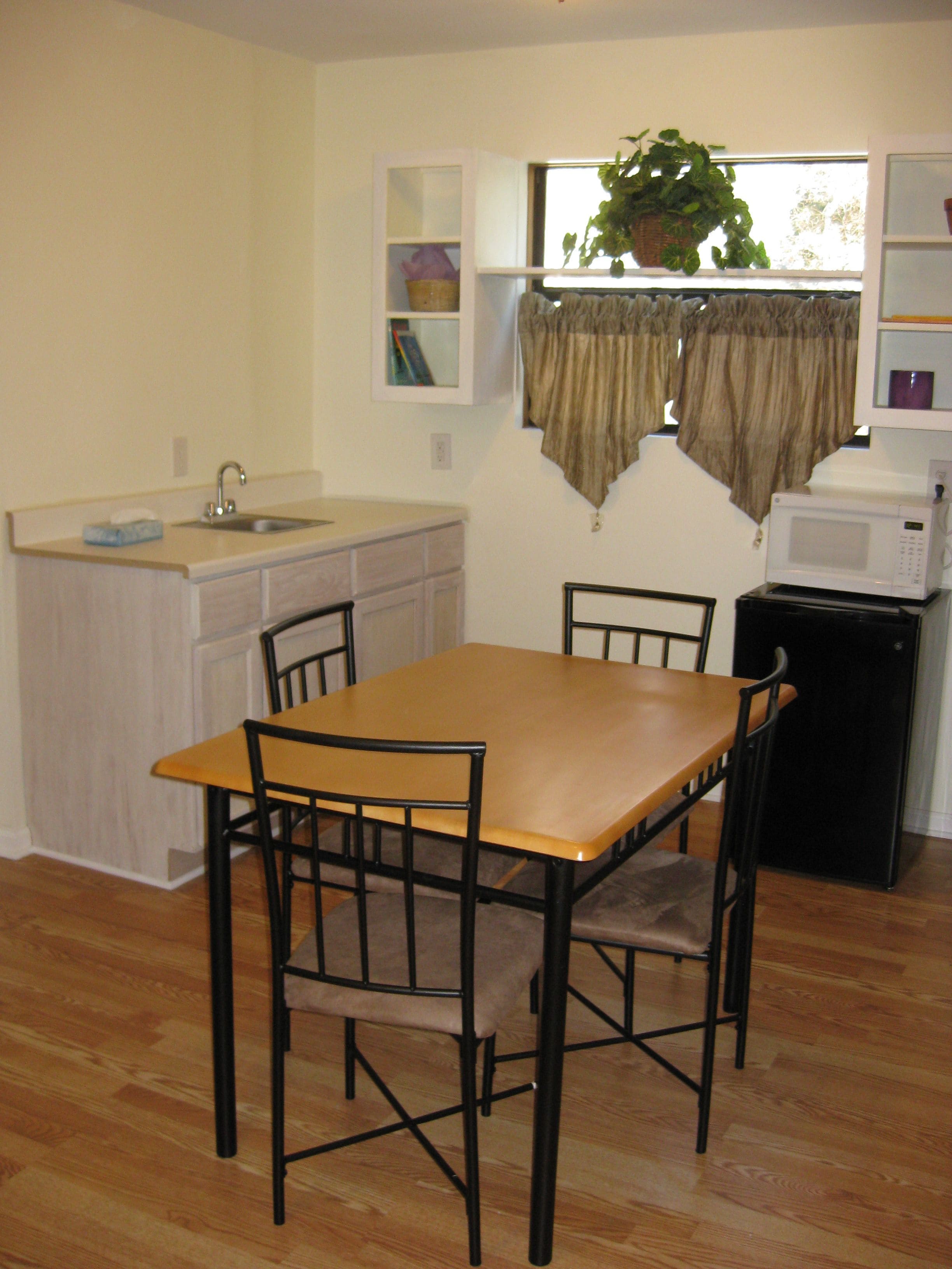 A compact kitchen with a wooden table and chairs, a built-in sink, a microwave, and a window with curtains.
