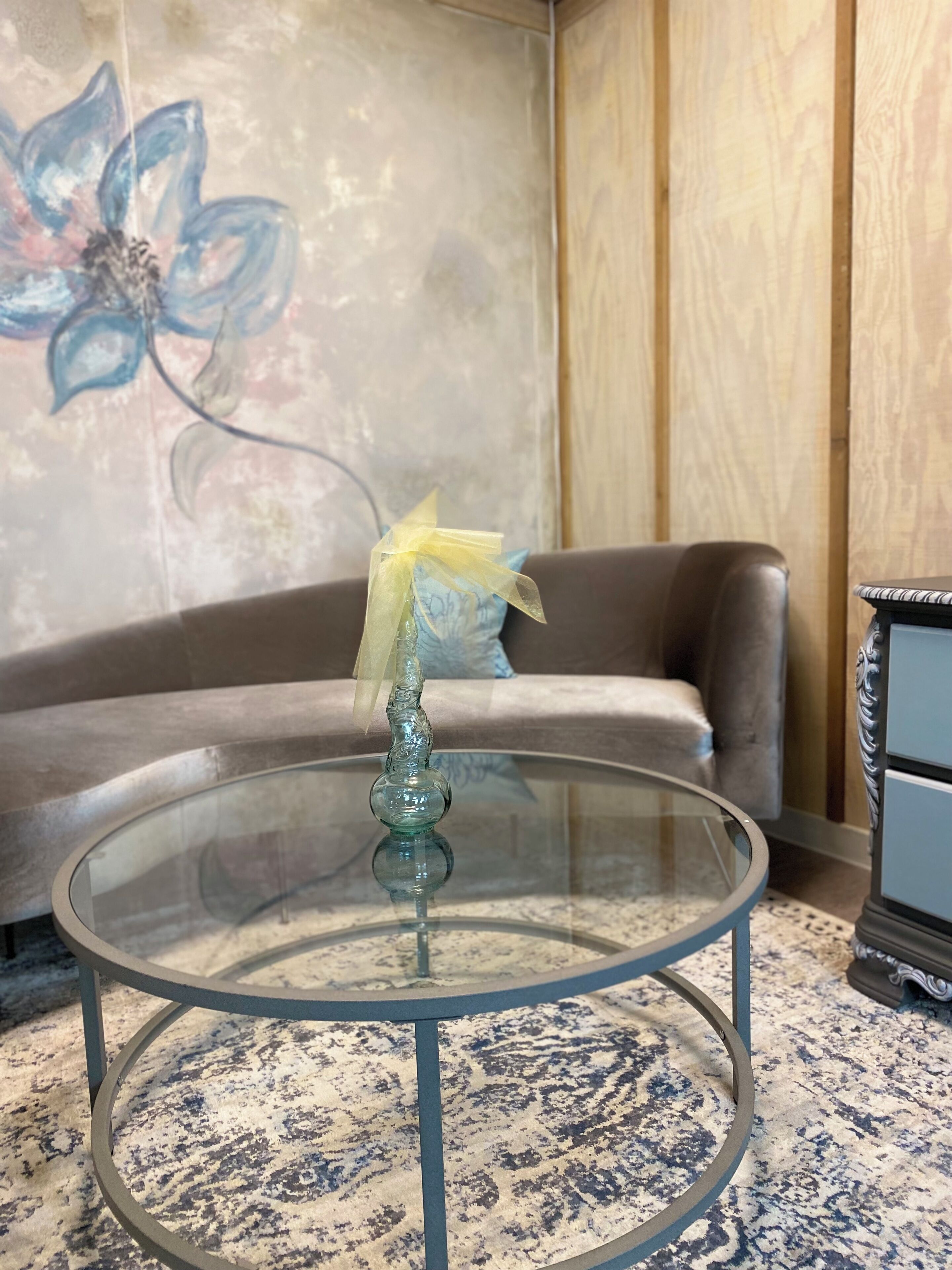Una mesa de centro redonda de vidrio con una base decorativa, un sofá marrón con un cojín y un armario de madera en una habitación con alfombra estampada.