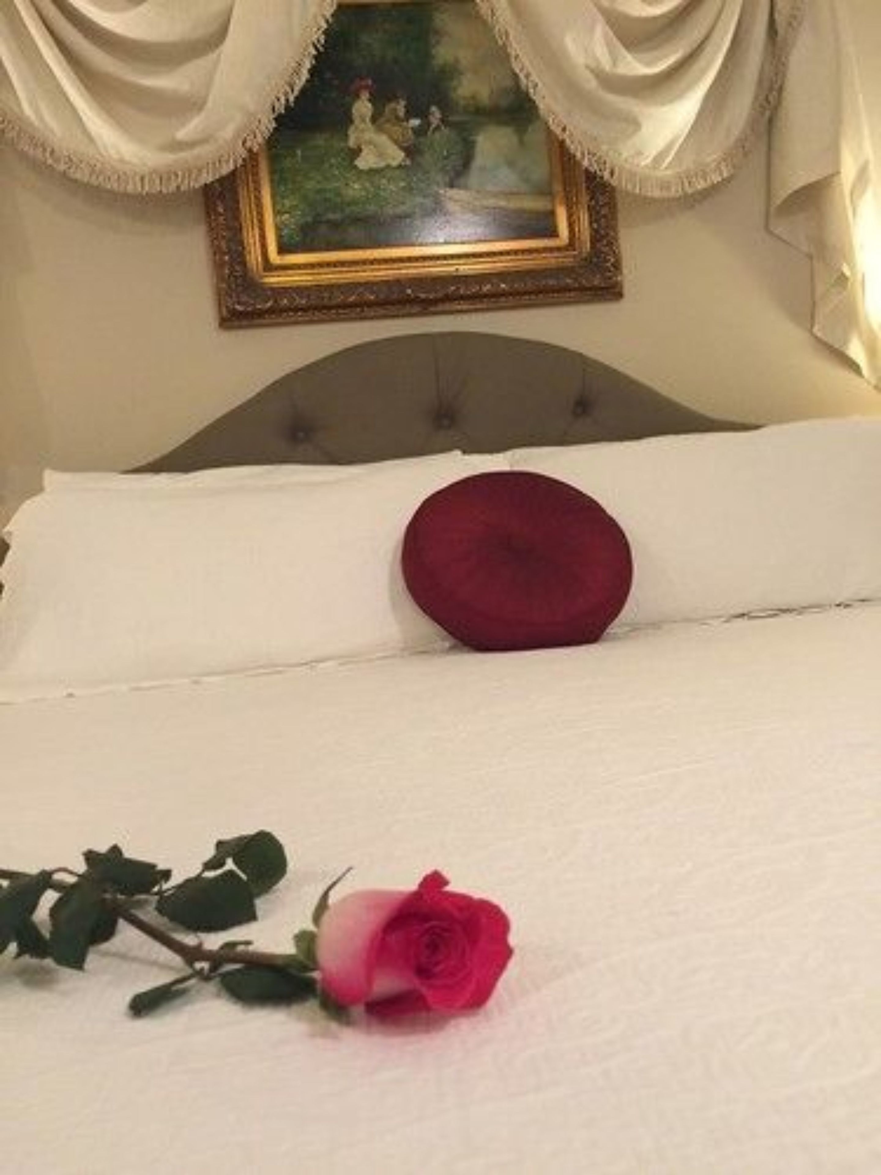Uma cama com um cobreleito branco, um travesseiro vermelho e uma única rosa rosa sobre a cama.