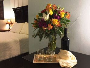 A hotel room with a bed, a bedside table with a vase of flowers, two wine glasses, and a bottle of wine.