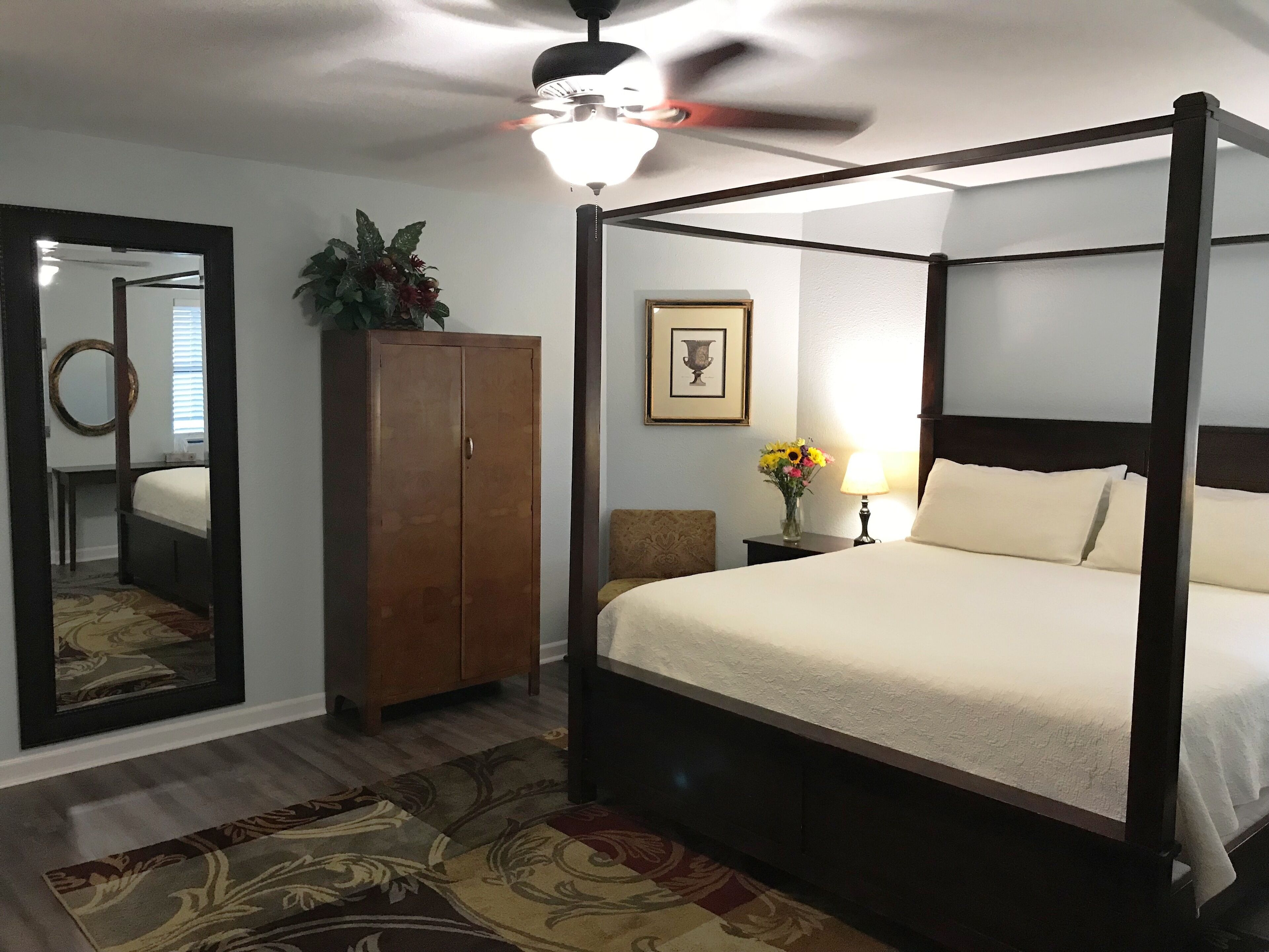 A four-poster bed with a nightstand, a mirror, a wardrobe, and a ceiling fan in a hotel room.