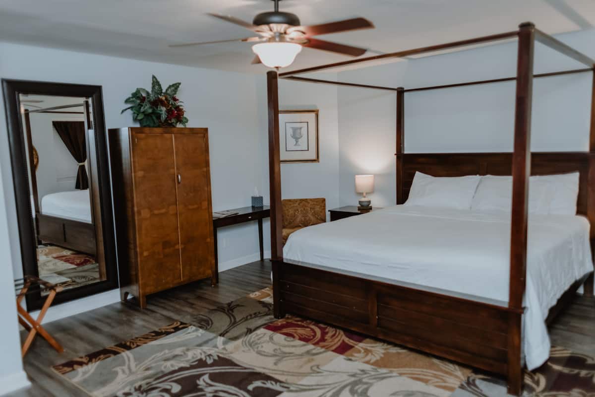 A four-poster bed with a wooden frame and white linens, a wooden wardrobe, a mirror, a ceiling fan, and a floral-patterned rug.