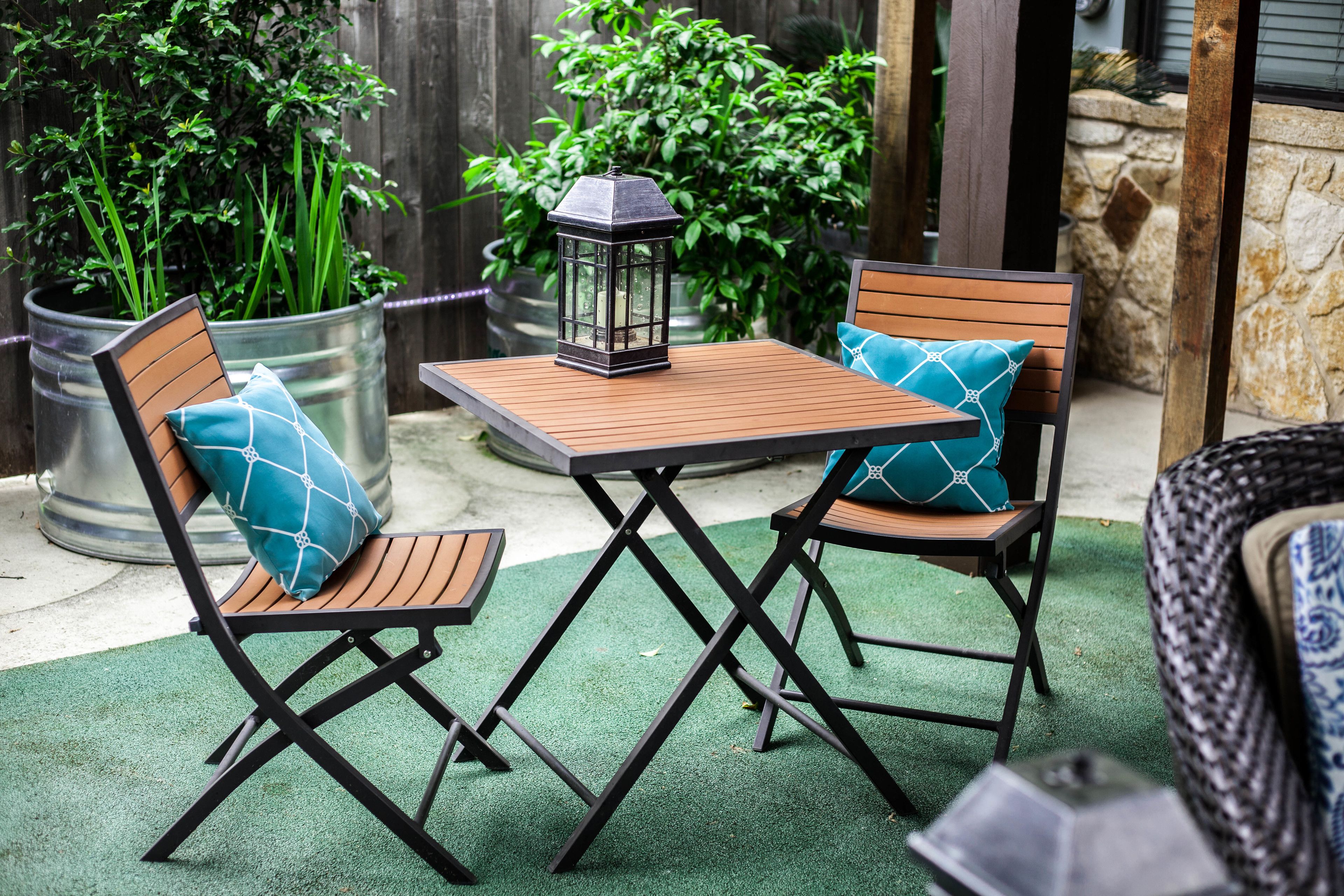 Un patio con dos sillas de madera y una mesa, rodeado de plantas en macetas y un muro de piedra.