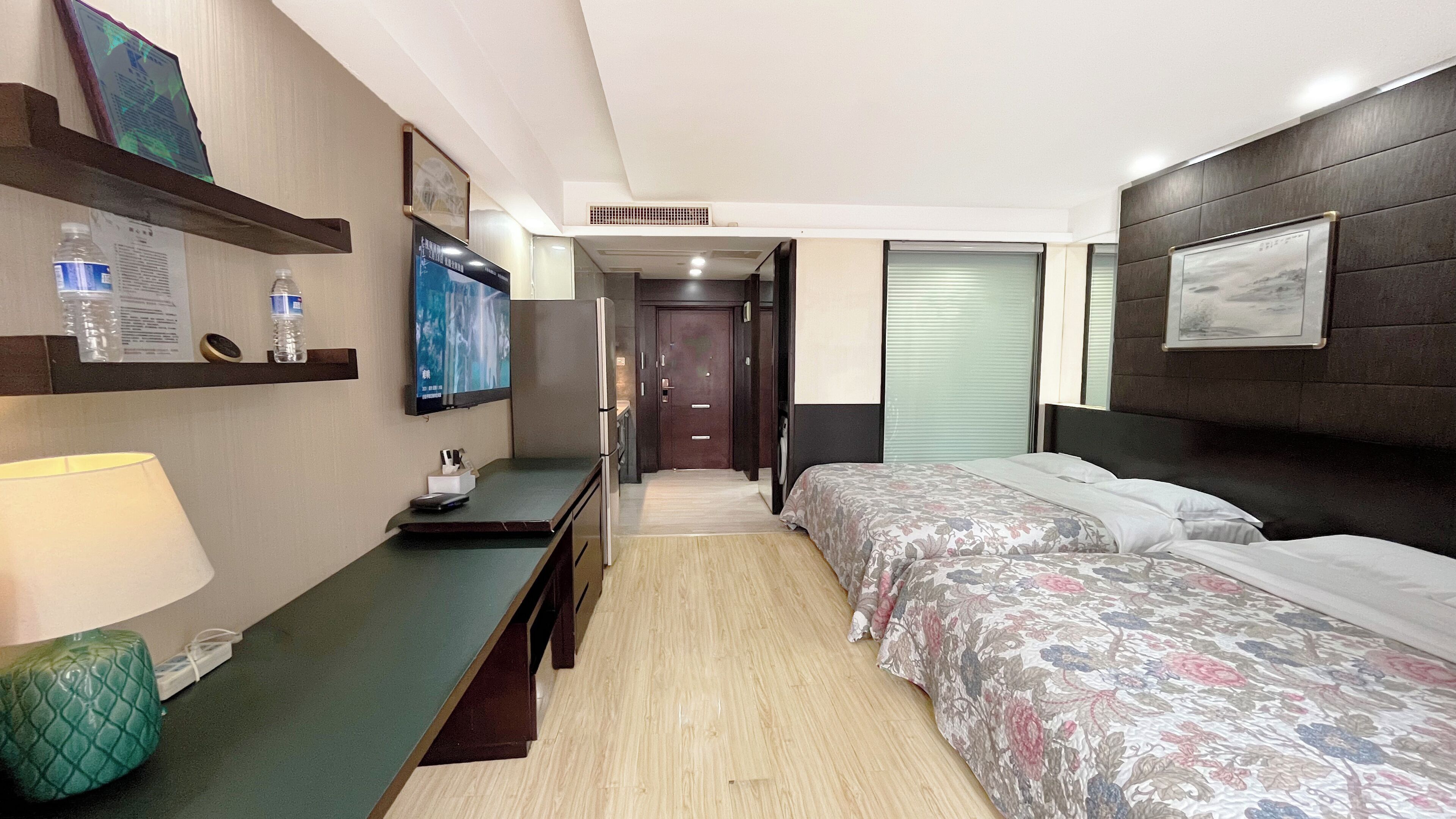 A hotel room with a bed, a desk with a lamp, a TV, and a door leading to another room.