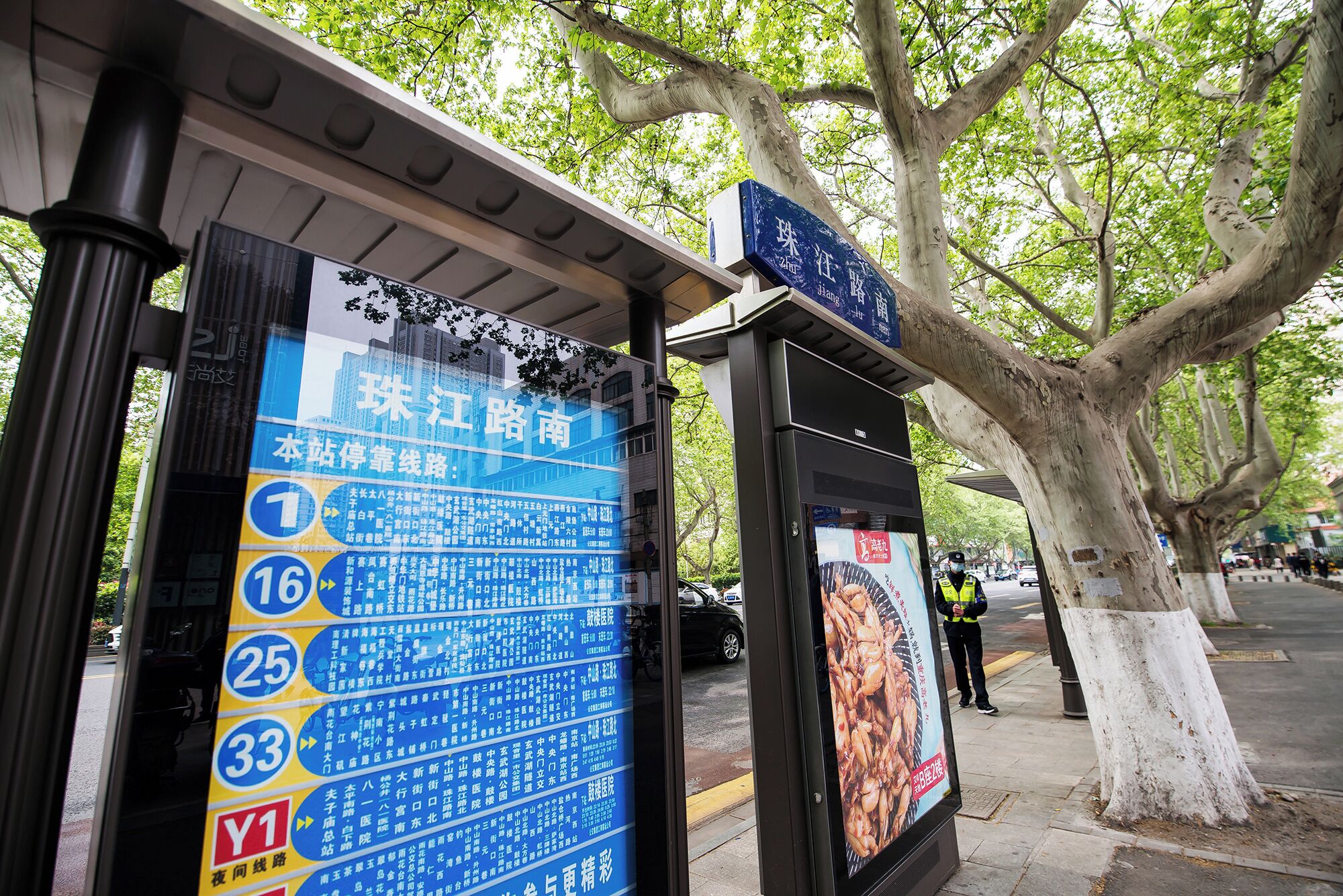 A bus stop shelter with a large blue sign listing bus routes and a smaller sign above it.