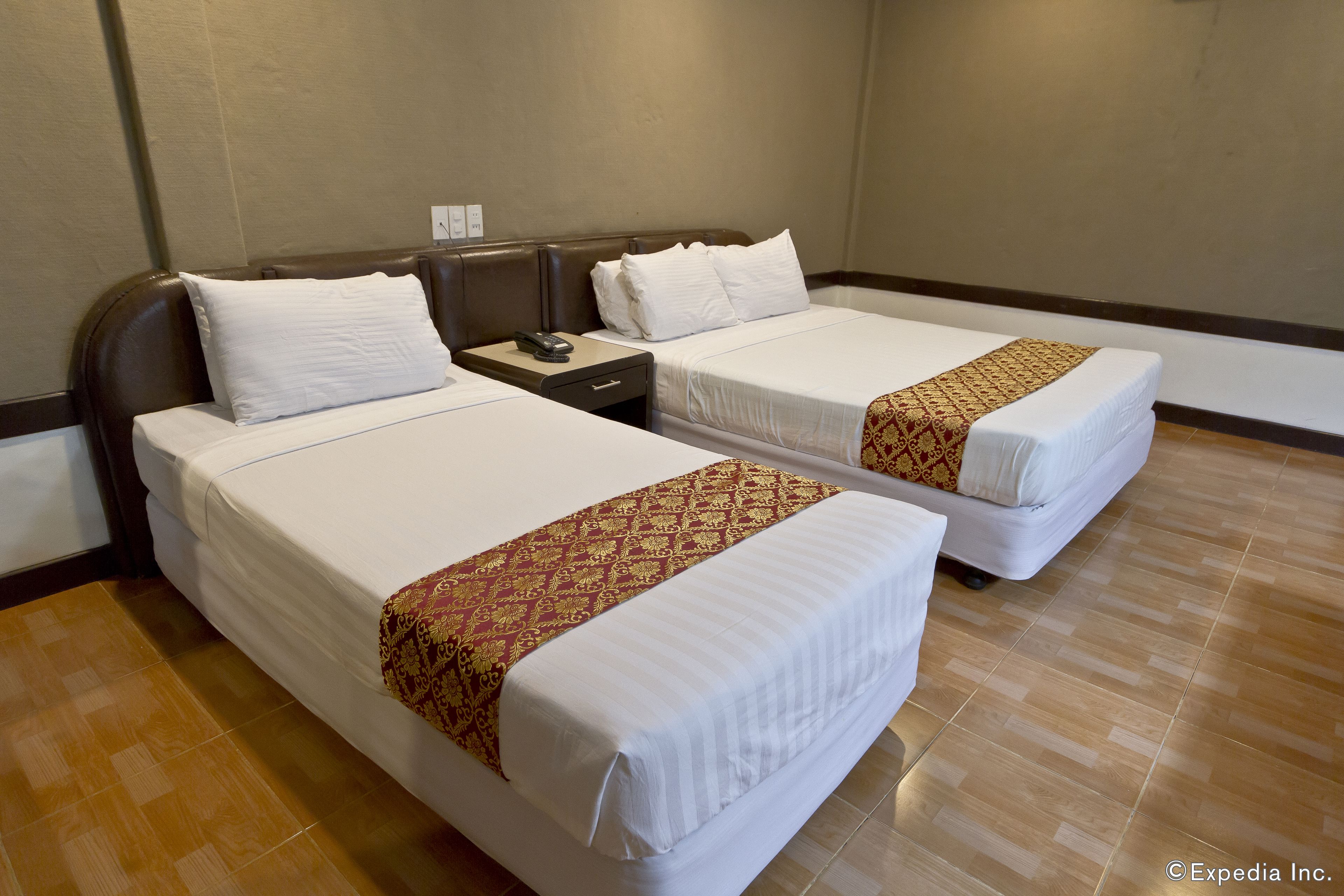 Two single beds with white linens and patterned bedspreads, a nightstand with a phone, and a plain wall with a power outlet.