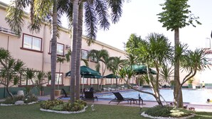 A pool area with lounge chairs and umbrellas, surrounded by palm trees and a building with a light-colored facade. - Holiday Spa Hotel (Cebu City)