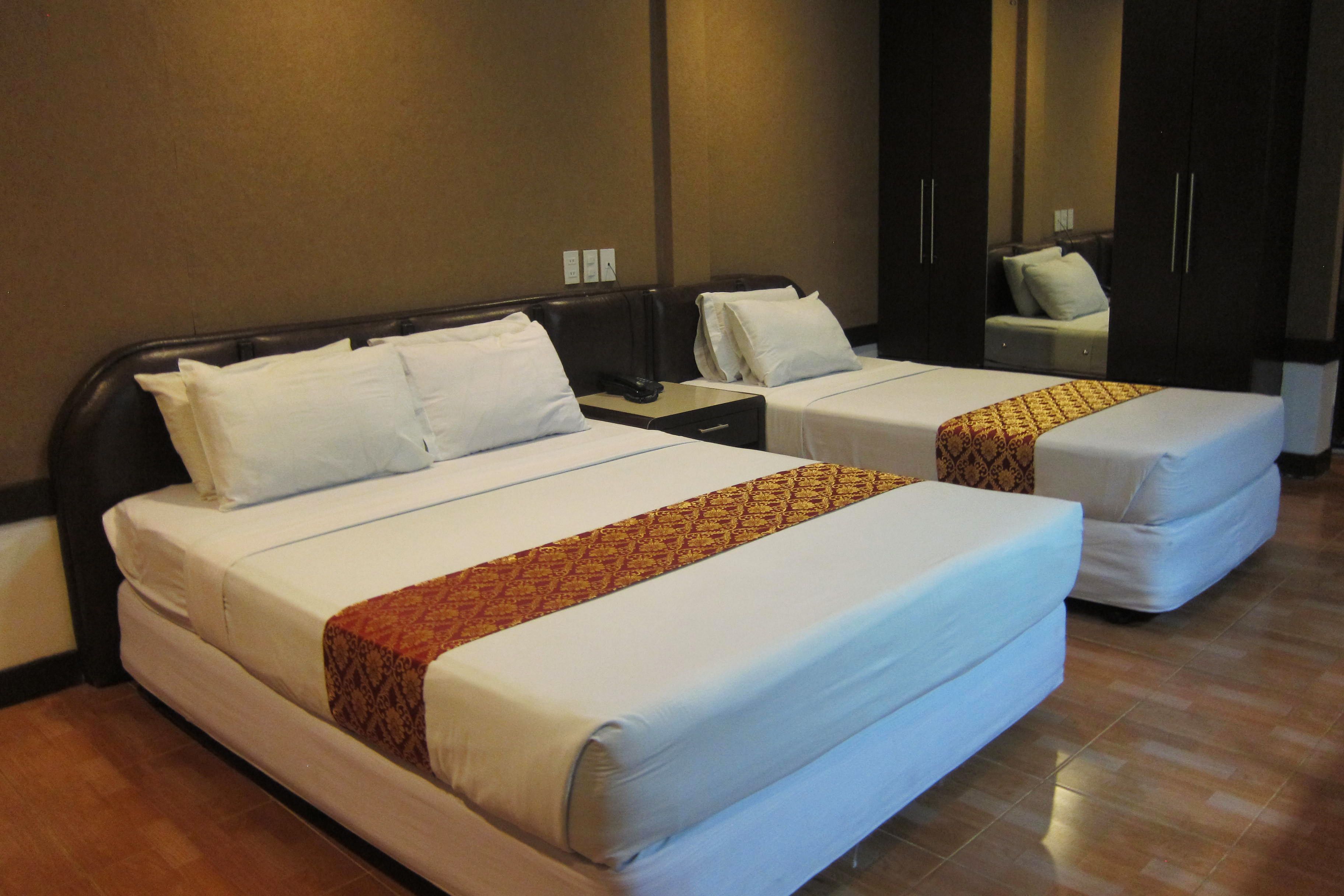 A hotel room with two single beds, each with white linens and a patterned throw. There are dark wooden wardrobes and a small bedside table with a lamp.