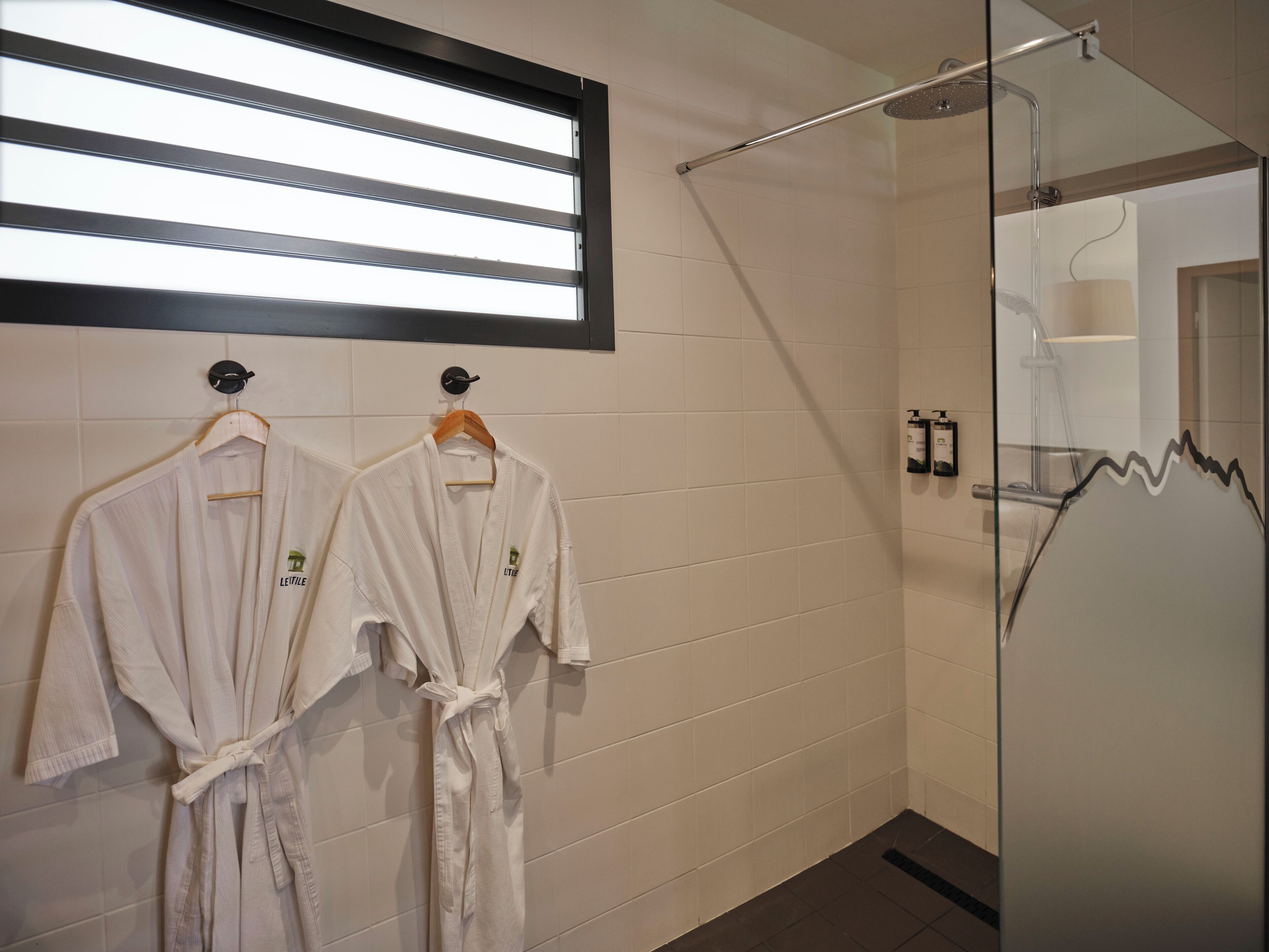 A bathroom with two white robes hanging on hooks, a glass shower enclosure, and a window with horizontal blinds.