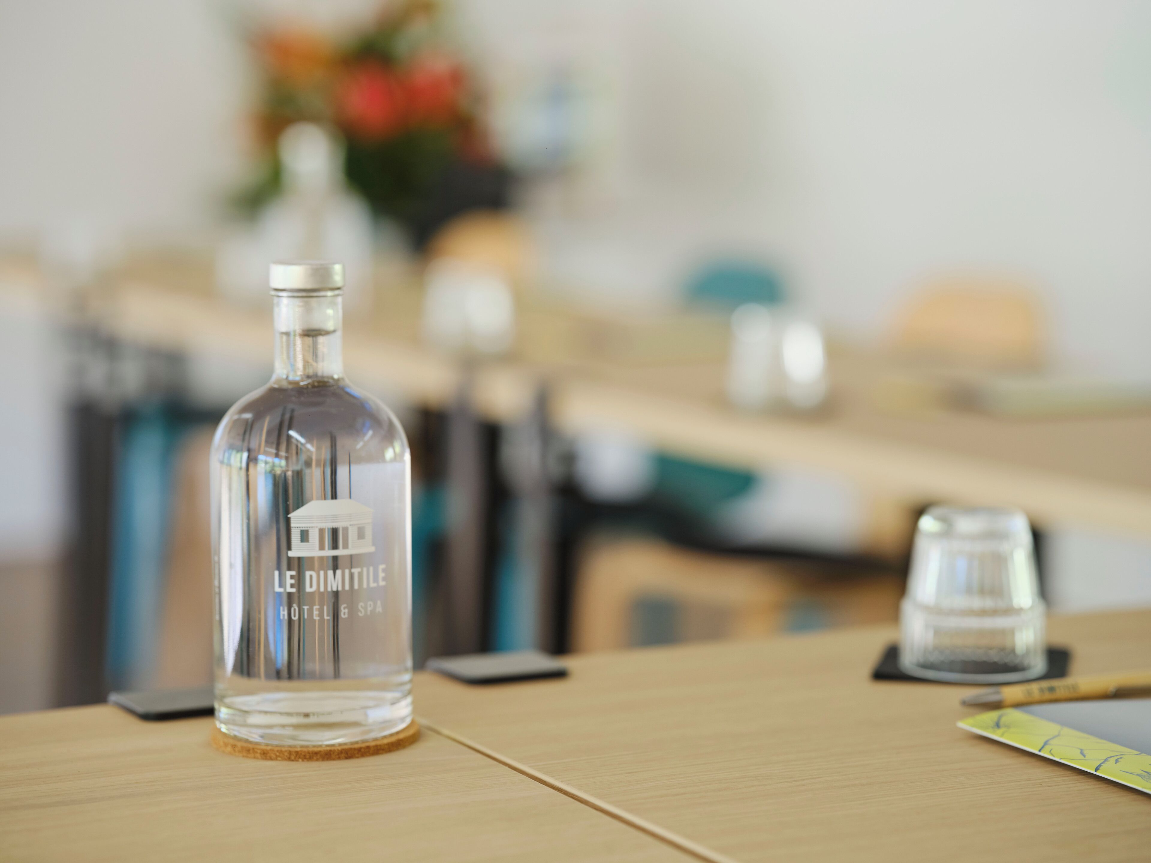 Una botella transparente con la etiqueta “LE DIMITLE HOTEL & SPA” sobre una mesa de madera, junto a un teléfono inteligente y un bloc de notas.