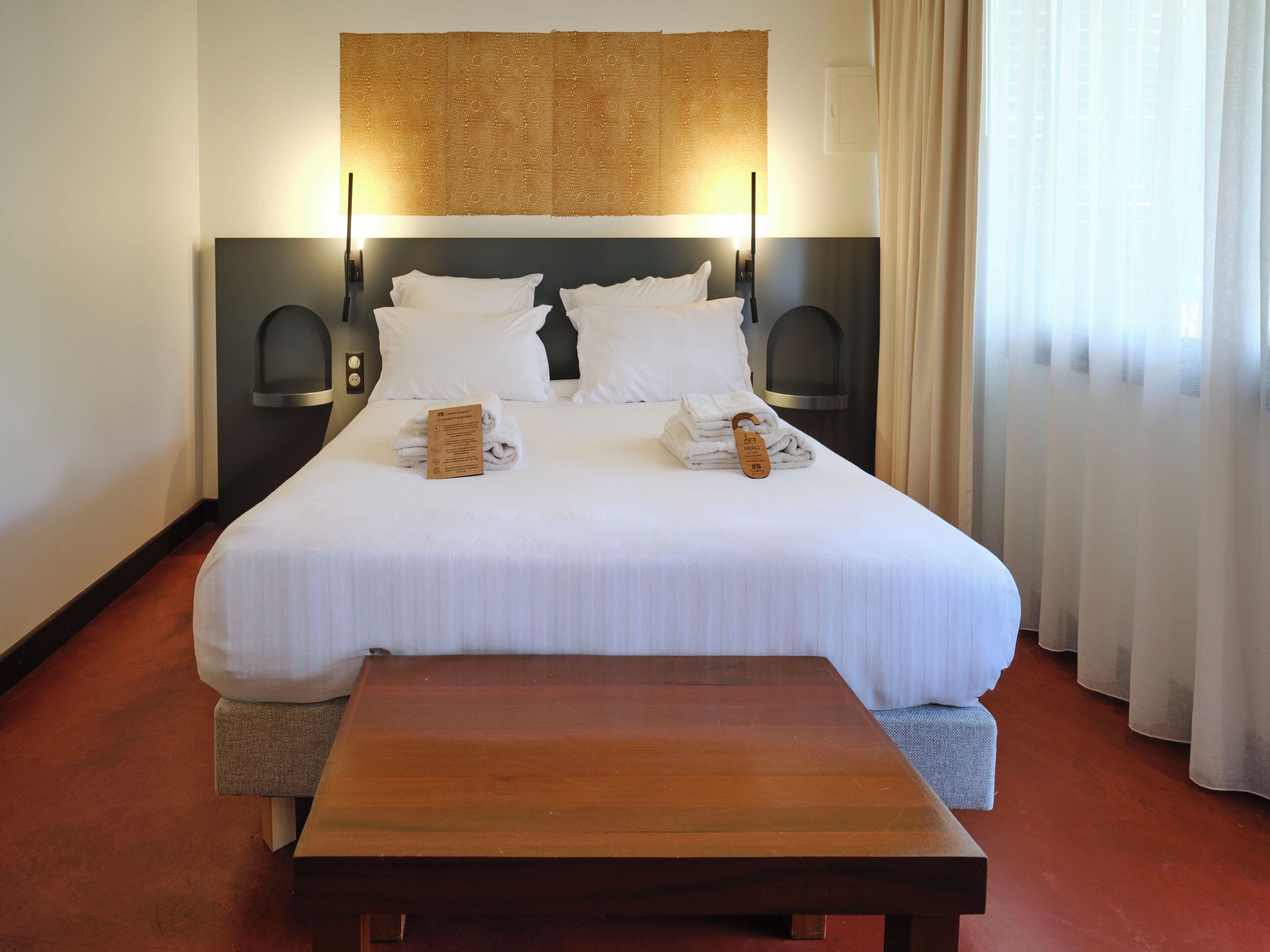 Una cama bien tendida con sábanas blancas, dos mesitas de noche, una mesita auxiliar de madera y un ventanal con cortinas translúcidas.