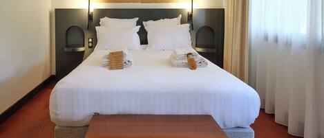 Een netjes opgemaakt bed met witte lakens, twee nachtkastjes, een houten nachtkastje, en een groot raam met dunne gordijnen.