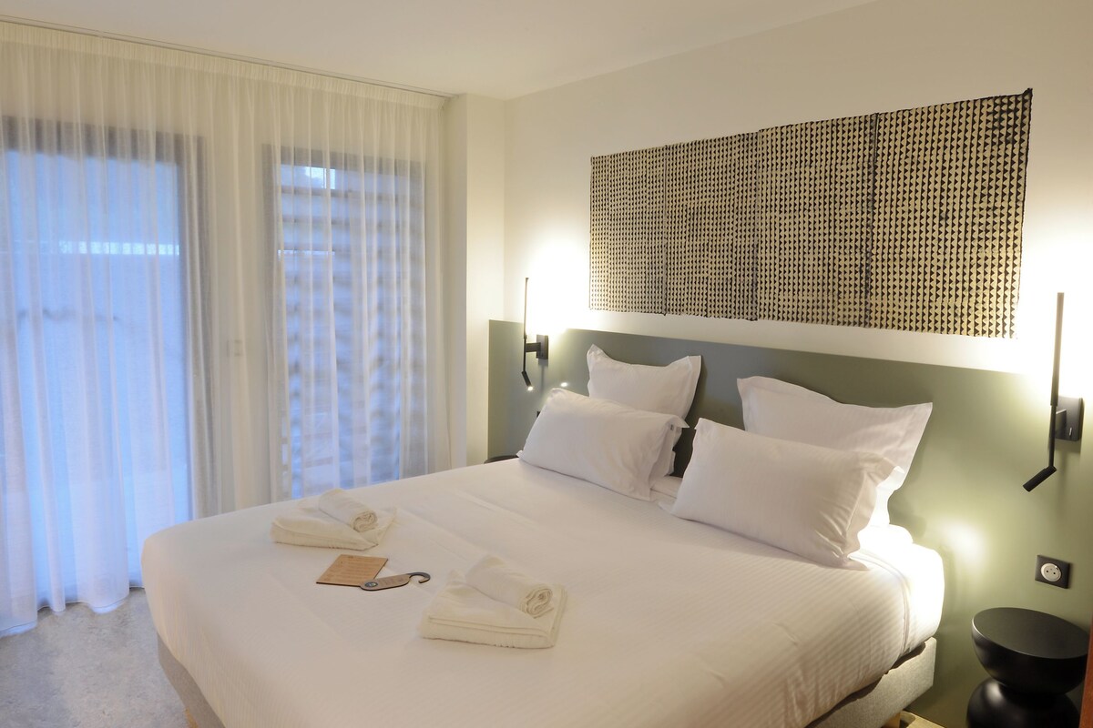 A hotel room with a bed, ceiling fan, curtains, wall-mounted light, and a textured wall decoration.