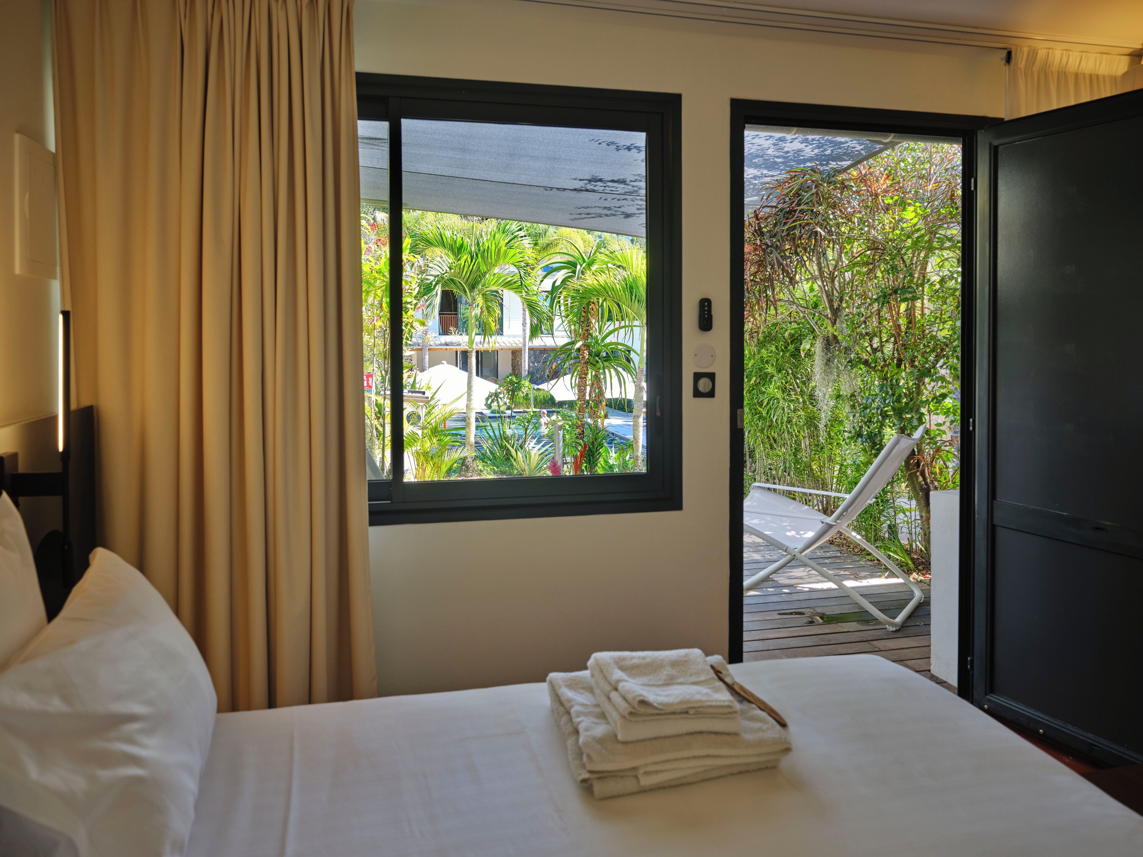 A hotel room with a bed, a window with a view of a pool and greenery, and a door leading to an outdoor area with a chair.