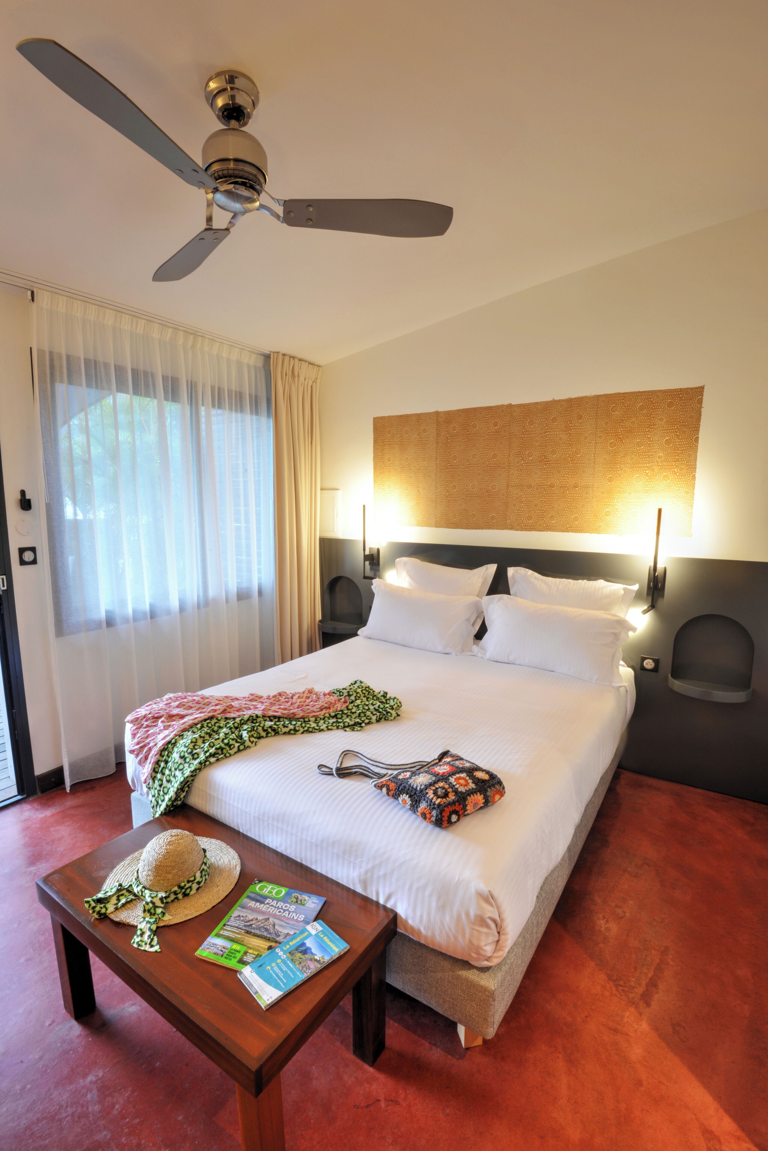 Una camera d'albergo con un letto, una ventola a soffitto, un tavolo con un cappello e riviste, e una finestra con tende.
