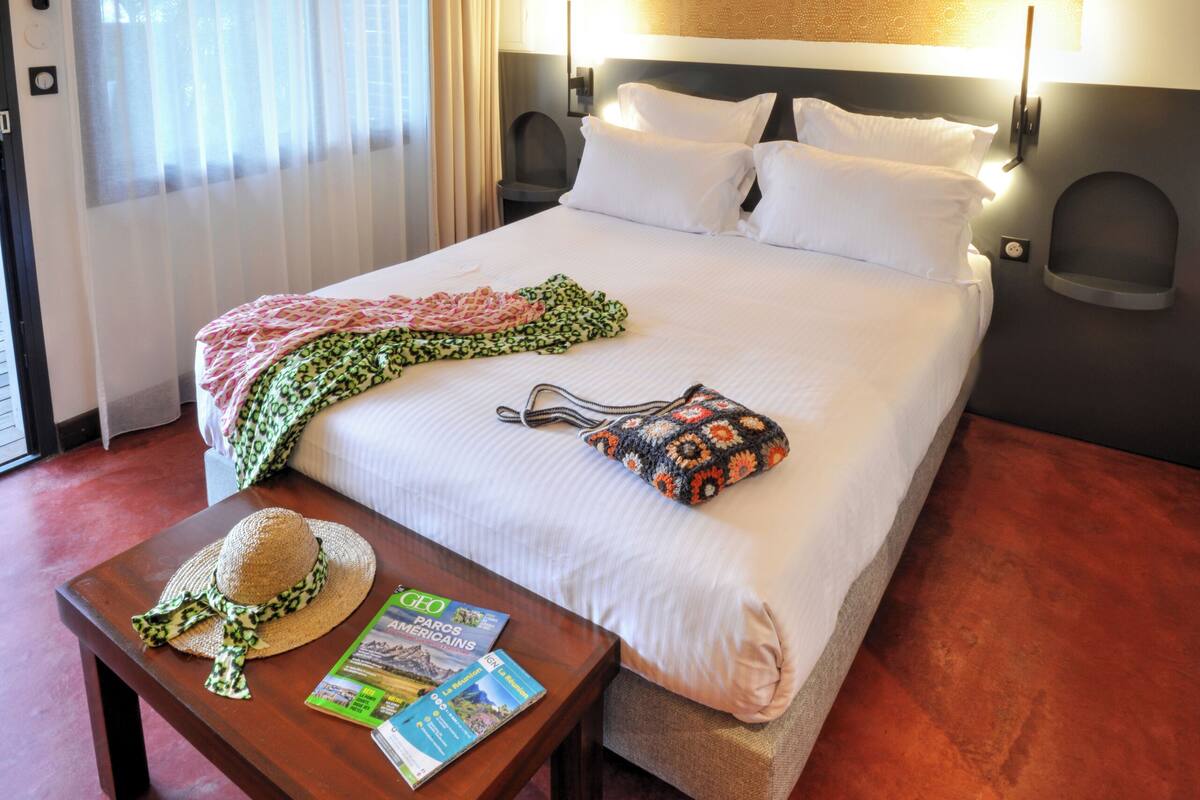 Quarto de hotel com cama, ventilador de teto, mesa com chapéu e revistas, e janela com cortinas.