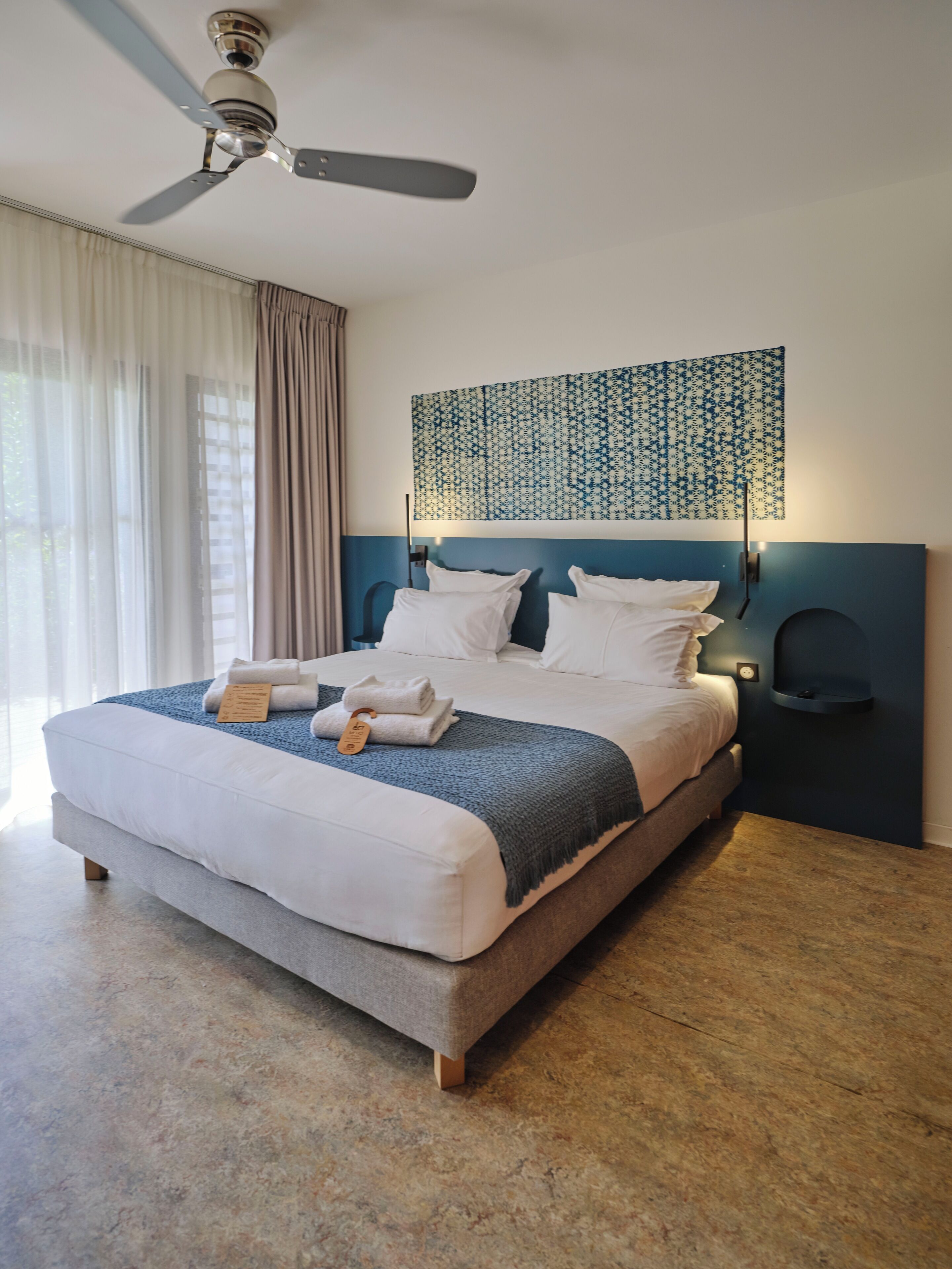A neatly made bed with white linens and a blue blanket, a ceiling fan, a wall-mounted light, and a patterned wall hanging.