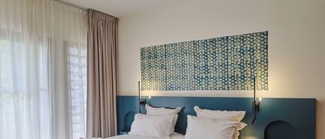 A neatly made bed with white linens and a blue blanket, a ceiling fan, a wall-mounted light, and a patterned wall hanging.