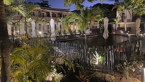 Vista nocturna de un patio de hotel con piscina, palmeras y zonas de estar al aire libre.