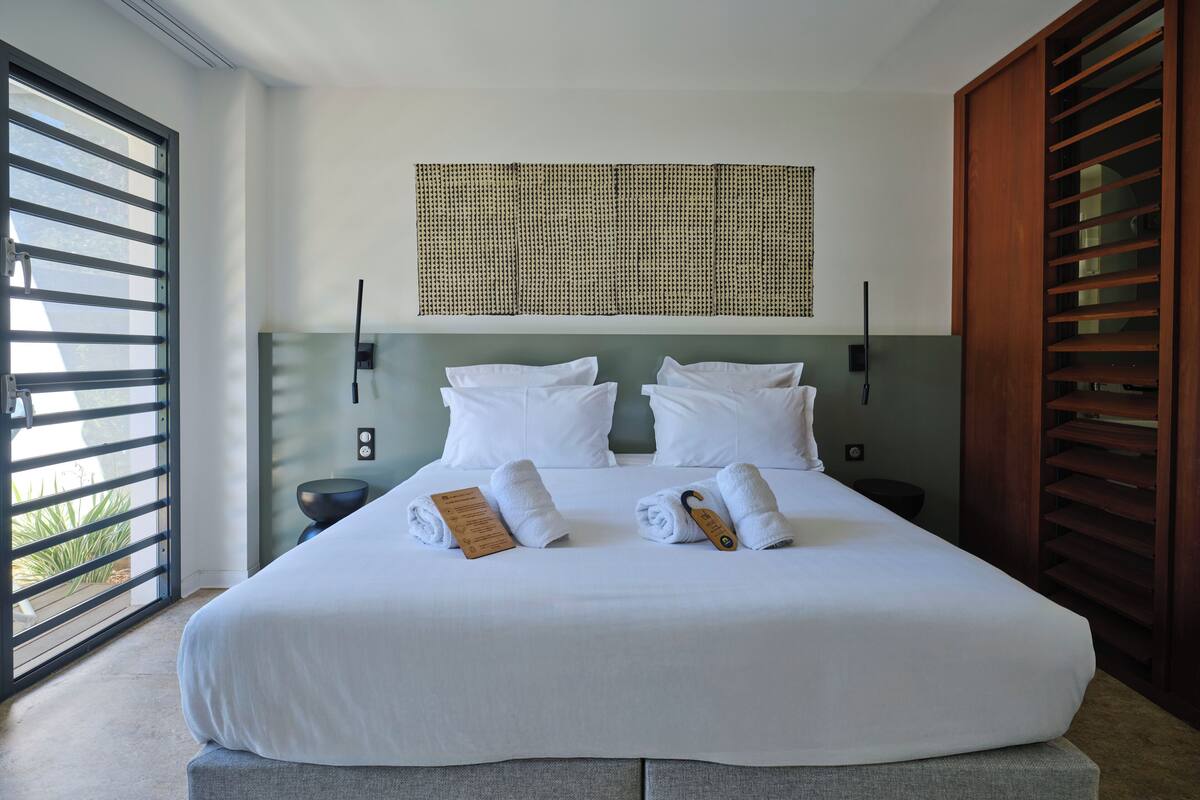Cama bem arrumada com roupa de cama branca, mesa de cabeceira, luminária fixada na parede e vista pela janela com persianas horizontais.