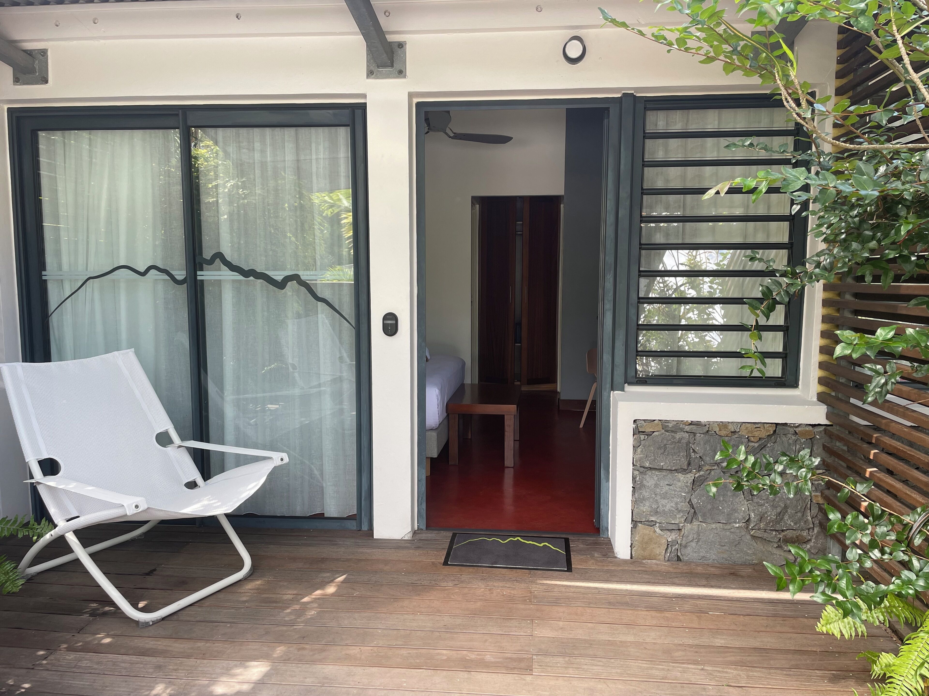 Um deck de madeira com uma cadeira branca, uma porta que dá para um quarto com cama e uma parede de pedra com janela veneziana de madeira.