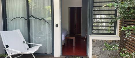 Una terraza de madera con una silla blanca, una puerta que da a una habitación con cama y un muro de piedra con ventana de listones de madera.