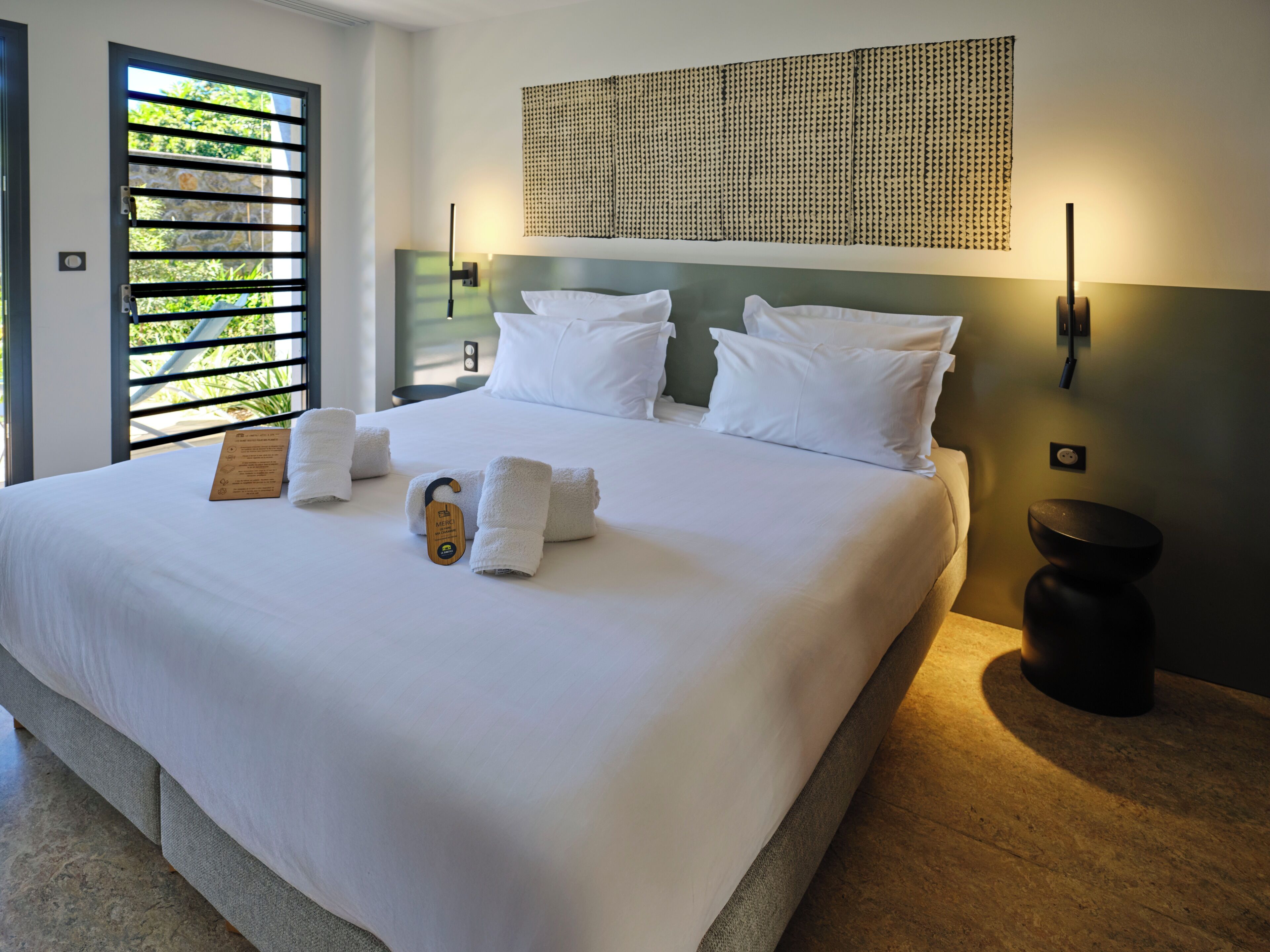 Habitación de hotel con una cama grande, ropa blanca, cabecera con panel tejido, mesita de noche con lámpara y vistas a zonas verdes a través de una ventana con persianas.