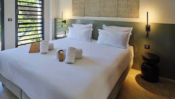 Habitación de hotel con una cama grande, ropa blanca, cabecera con panel tejido, mesita de noche con lámpara y vistas a zonas verdes a través de una ventana con persianas.