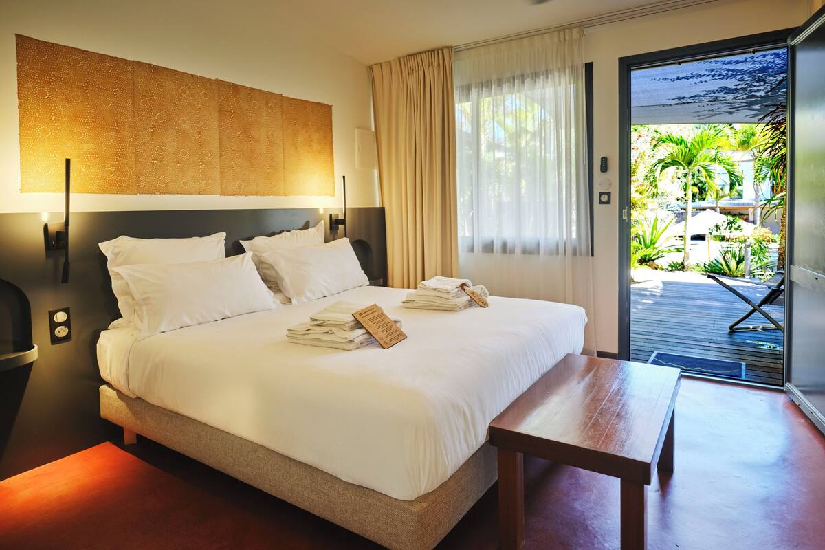 Quarto de hotel com uma cama grande, um banco de madeira, uma luminária fixada na parede e vista para um jardim e piscina pela janela.