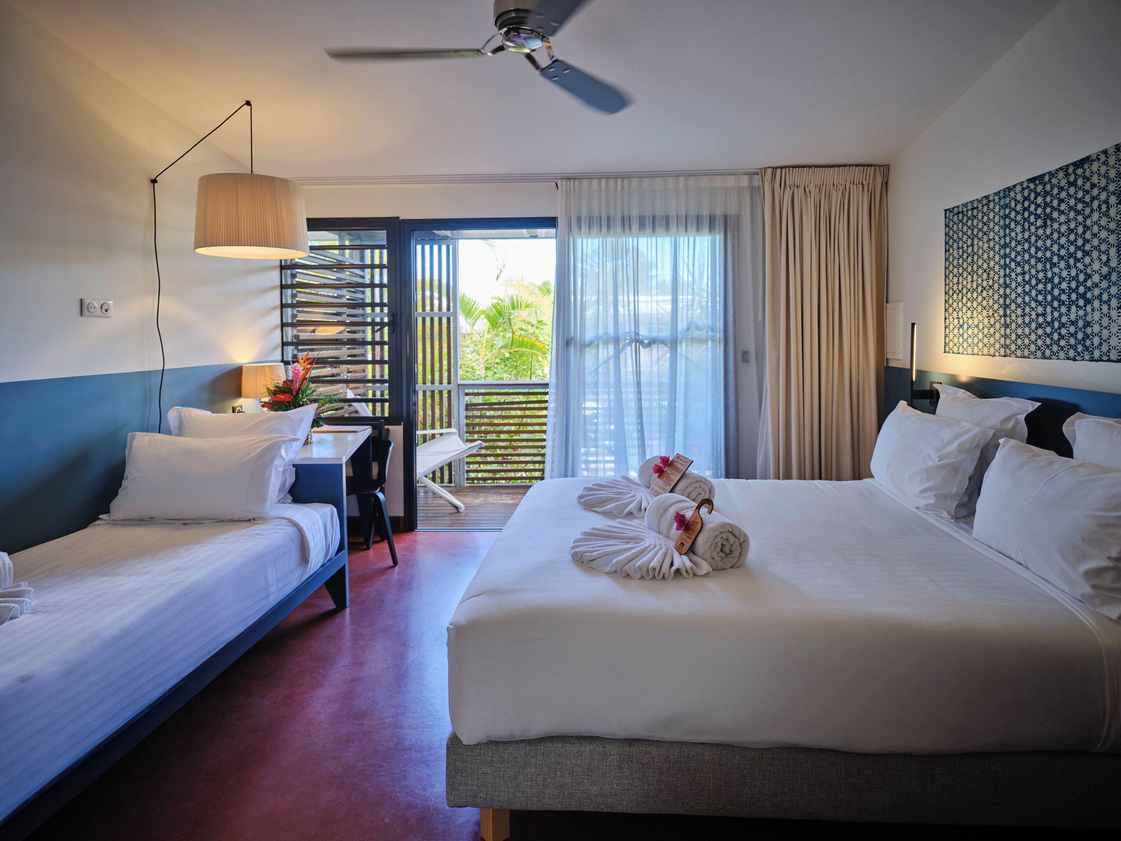 A hotel room with two beds, a desk, a chair, a ceiling fan, and a view of the outdoors.
