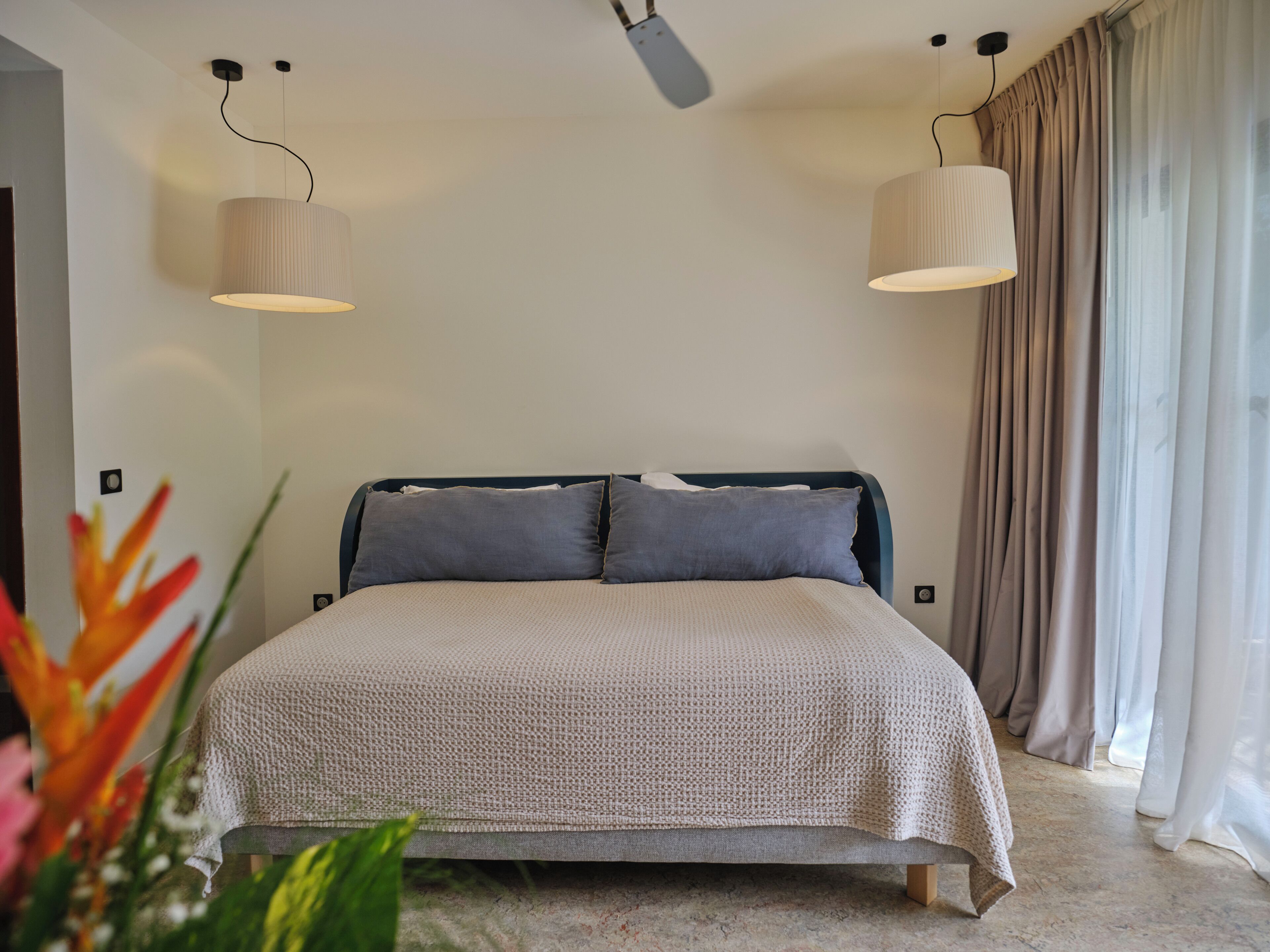A bedroom with a bed, two pendant lights, a window with curtains, and a plant in the foreground.