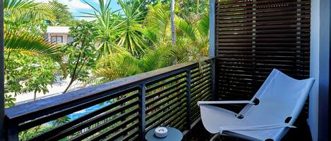 A balcony with a white chair, a small round table, and a view of a beach and palm trees.