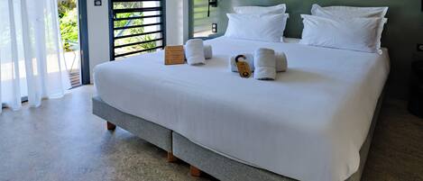 A modern hotel room with a large bed, white linens, and a view of greenery through the door.
