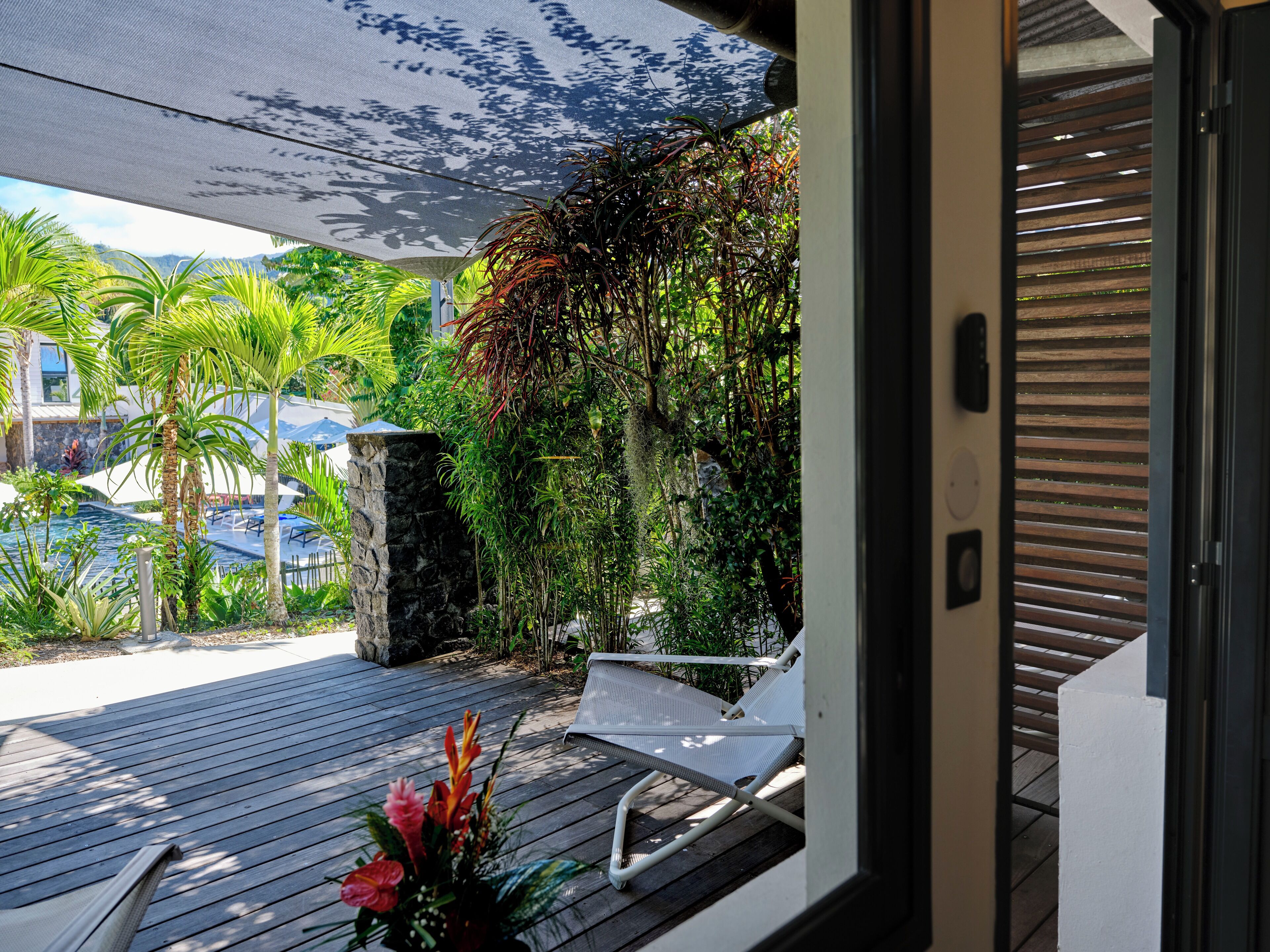 A wooden deck with a white chair and a hammock, surrounded by lush greenery and a view of a swimming pool.