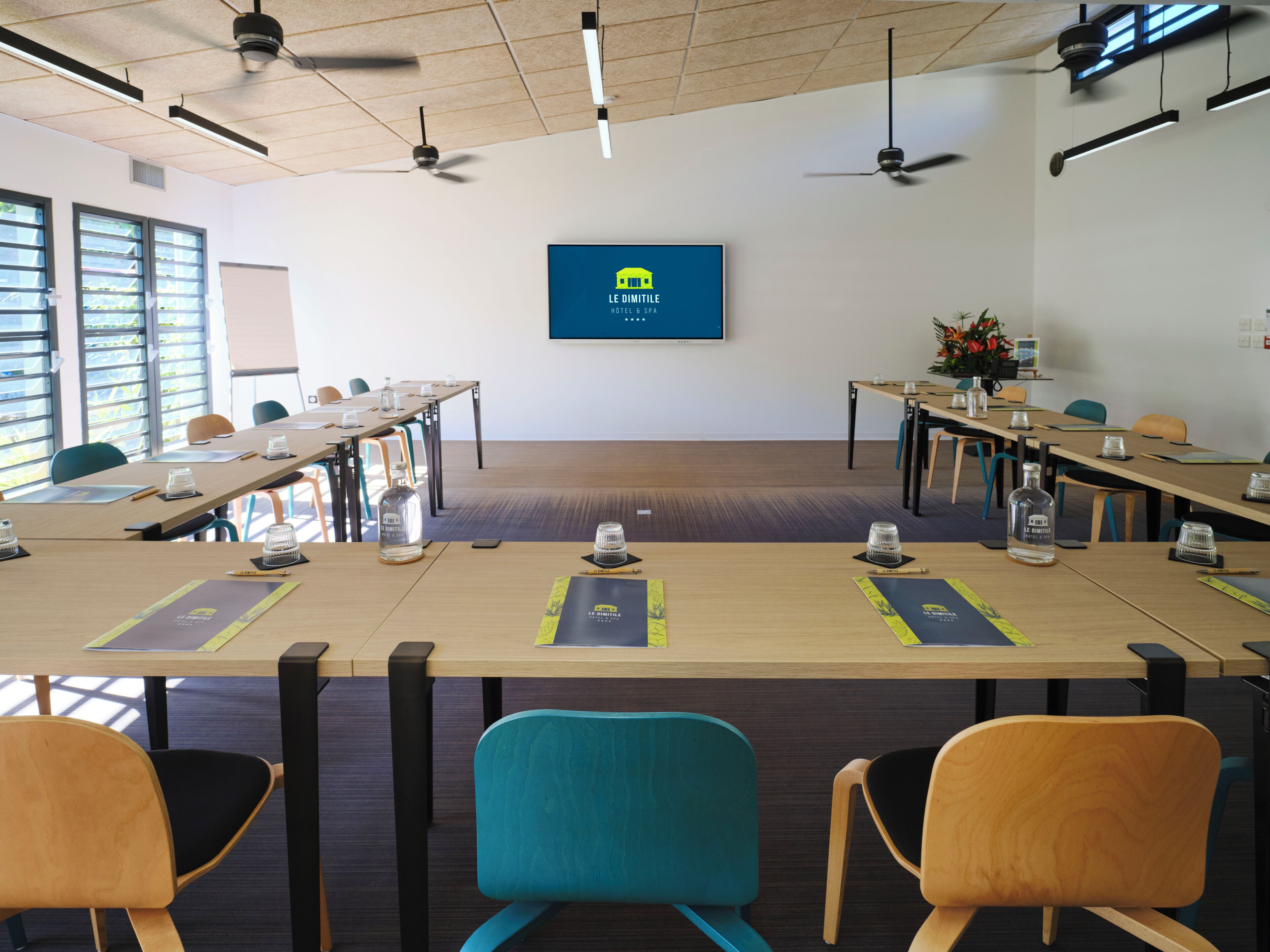 Une salle de conférence dotée d’un grand écran affichant le nom de l’hôtel, de plusieurs tables avec des chaises et de ventilateurs de plafond.